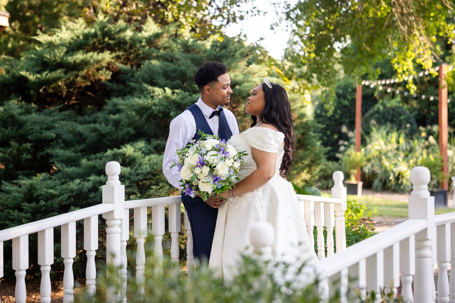 A bride and groom share a romantic moment on white stairs surrounded by lush greenery at an outdoor wedding venue.