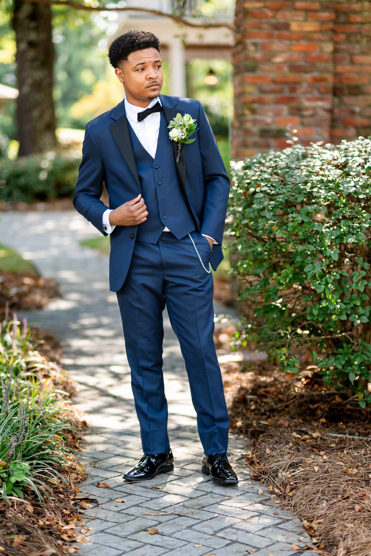 Young man in navy blue tuxedo with bow tie and boutonniere poses on brick pathway surrounded by lush greenery.