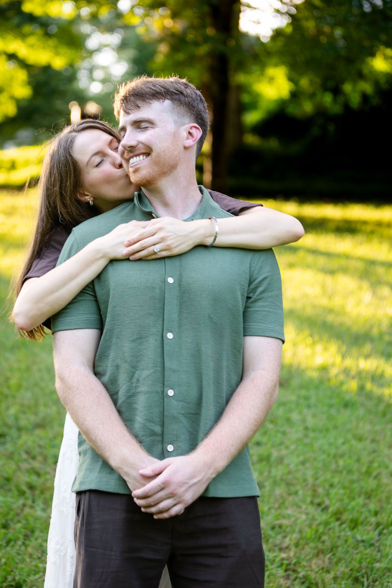 A woman hugs a smiling red-haired man from behind and kisses his cheek in a sunny green park.