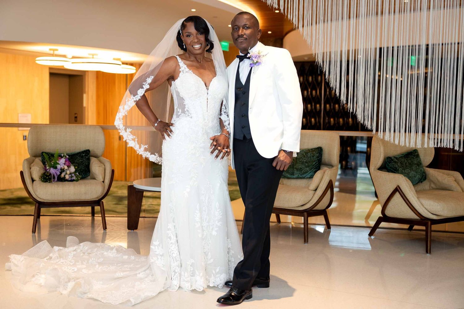 Elegant bride and groom posing in wedding attire inside a modern hotel lobby, bride wearing lace gown with veil.