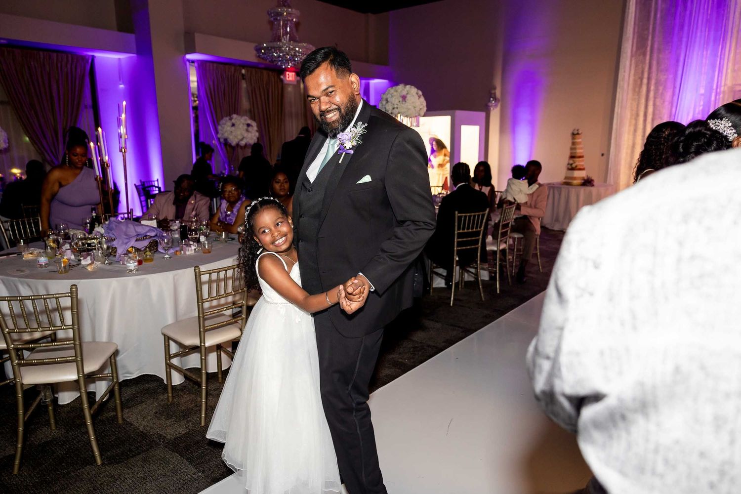Father dances with young flower girl in white dress at elegant wedding reception.