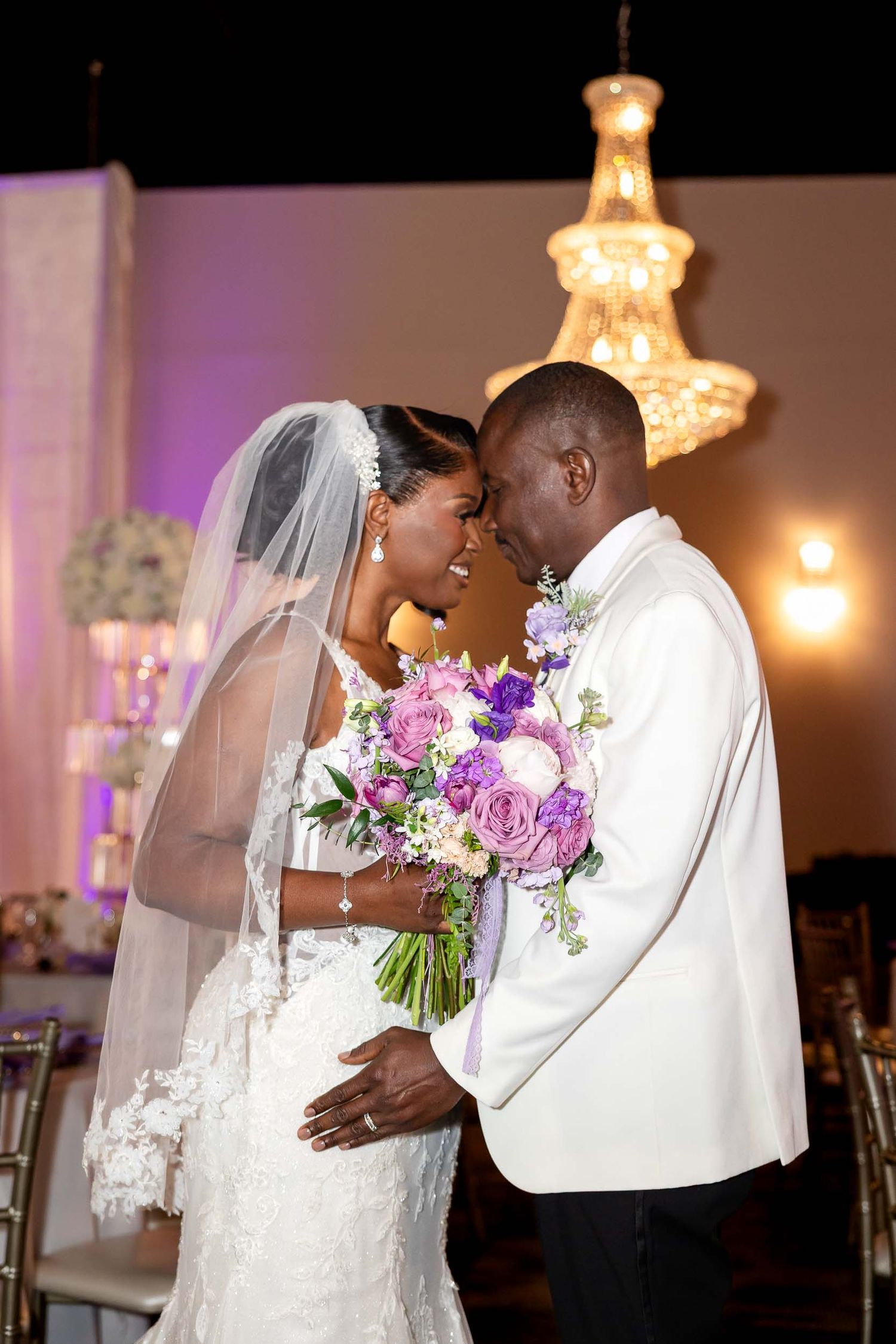 Bride and groom share romantic forehead touch holding purple bouquet under chandelier at elegant wedding reception.