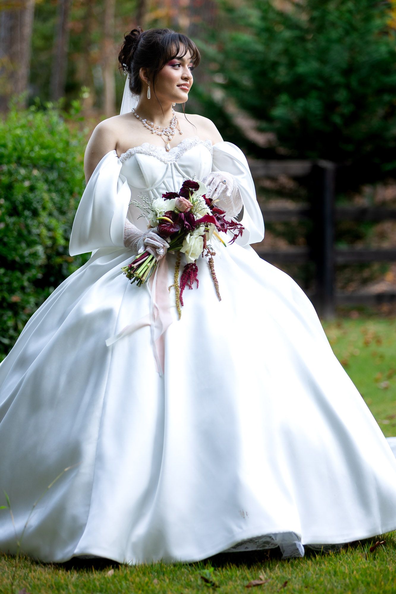 Bride in white ballgown holding burgundy bouquet outdoors among green trees.