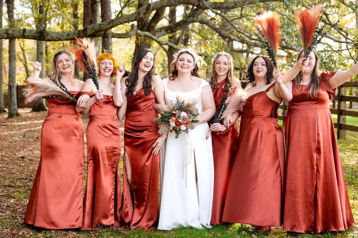 Bride in white gown with six bridesmaids in rust orange satin dresses holding feather bouquets outdoors in autumn setting.