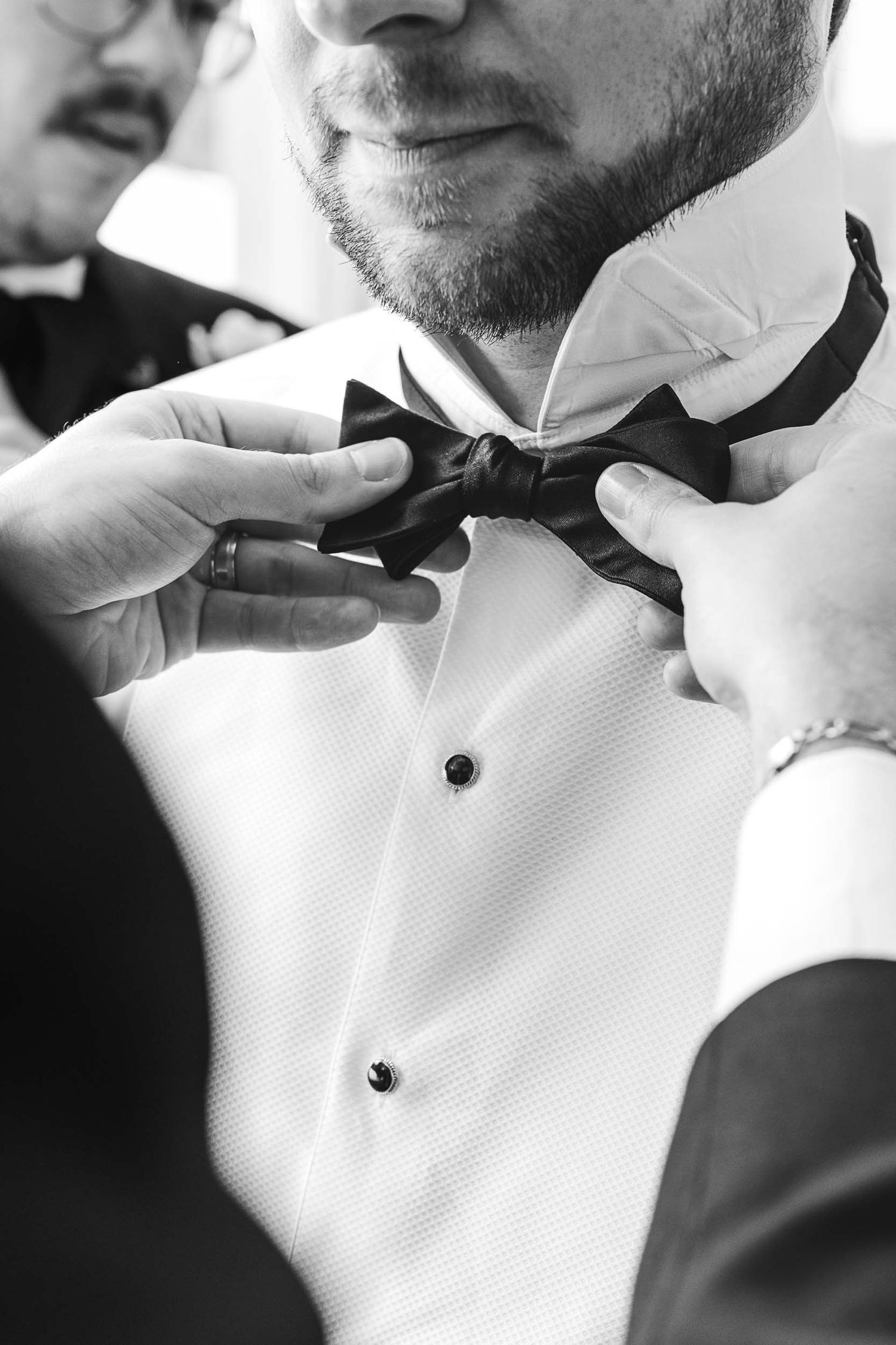 Hands adjusting a black bow tie on a groom wearing a white tuxedo shirt, black and white wedding photo.