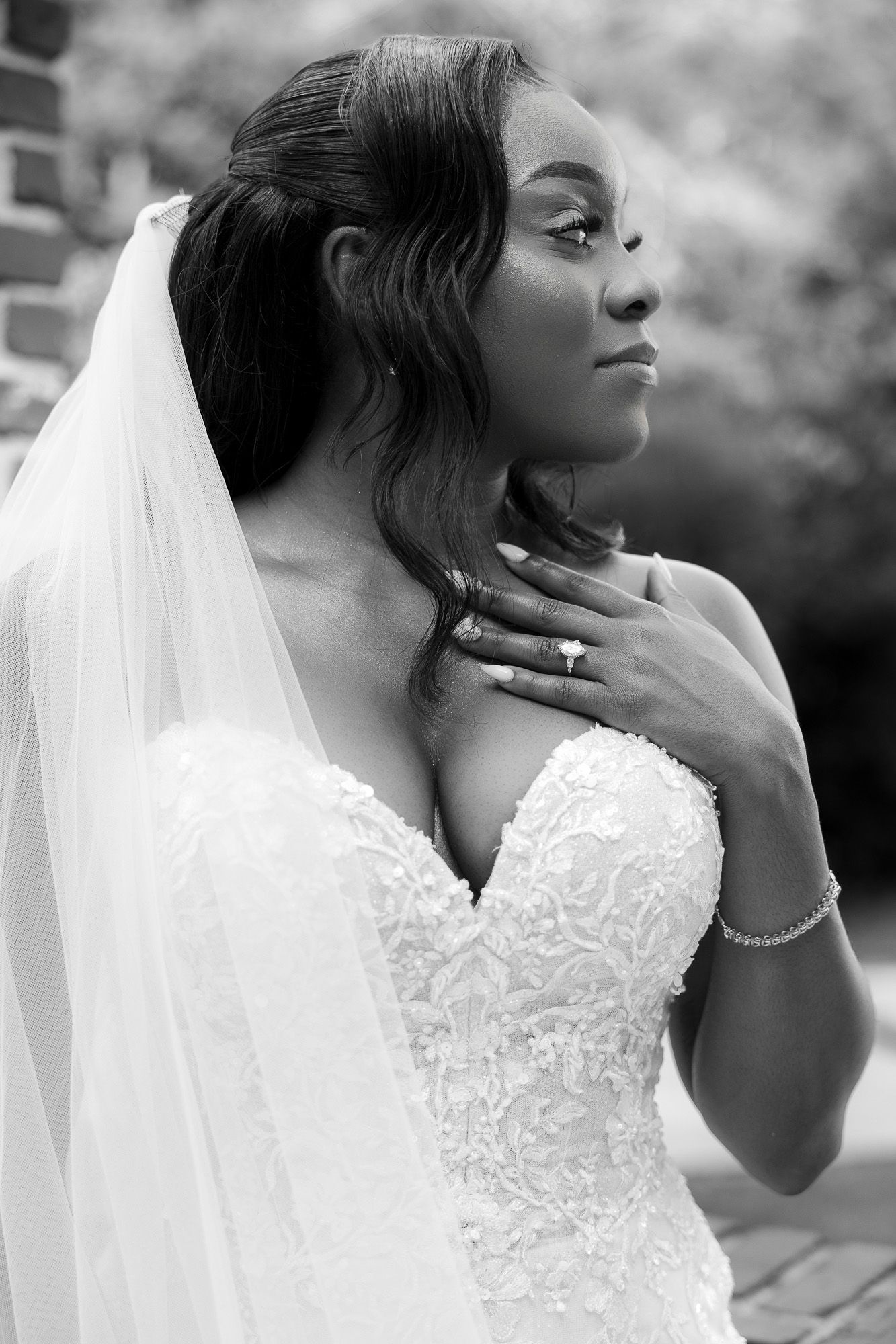 Black and white portrait of a bride in lace gown and veil, gracefully touching her neck with manicured hand.