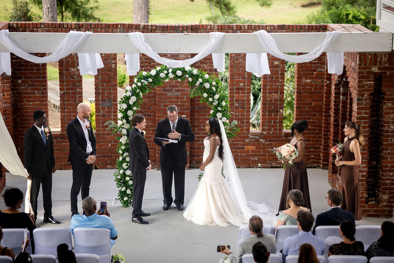 Outdoor wedding ceremony with bride and groom at floral arch before officiant, guests seated, brick wall backdrop.