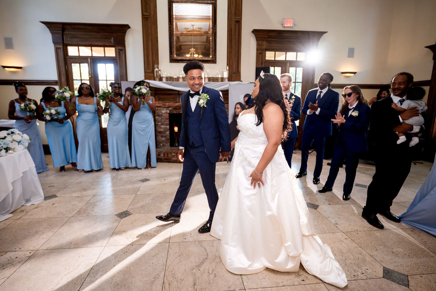 Bride and groom share first dance at wedding reception with bridesmaids in blue dresses and groomsmen in navy suits.