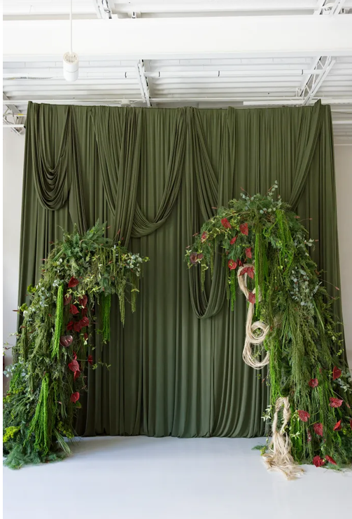 Elegant olive green draped fabric backdrop with lush greenery and red floral arrangements for a wedding ceremony.