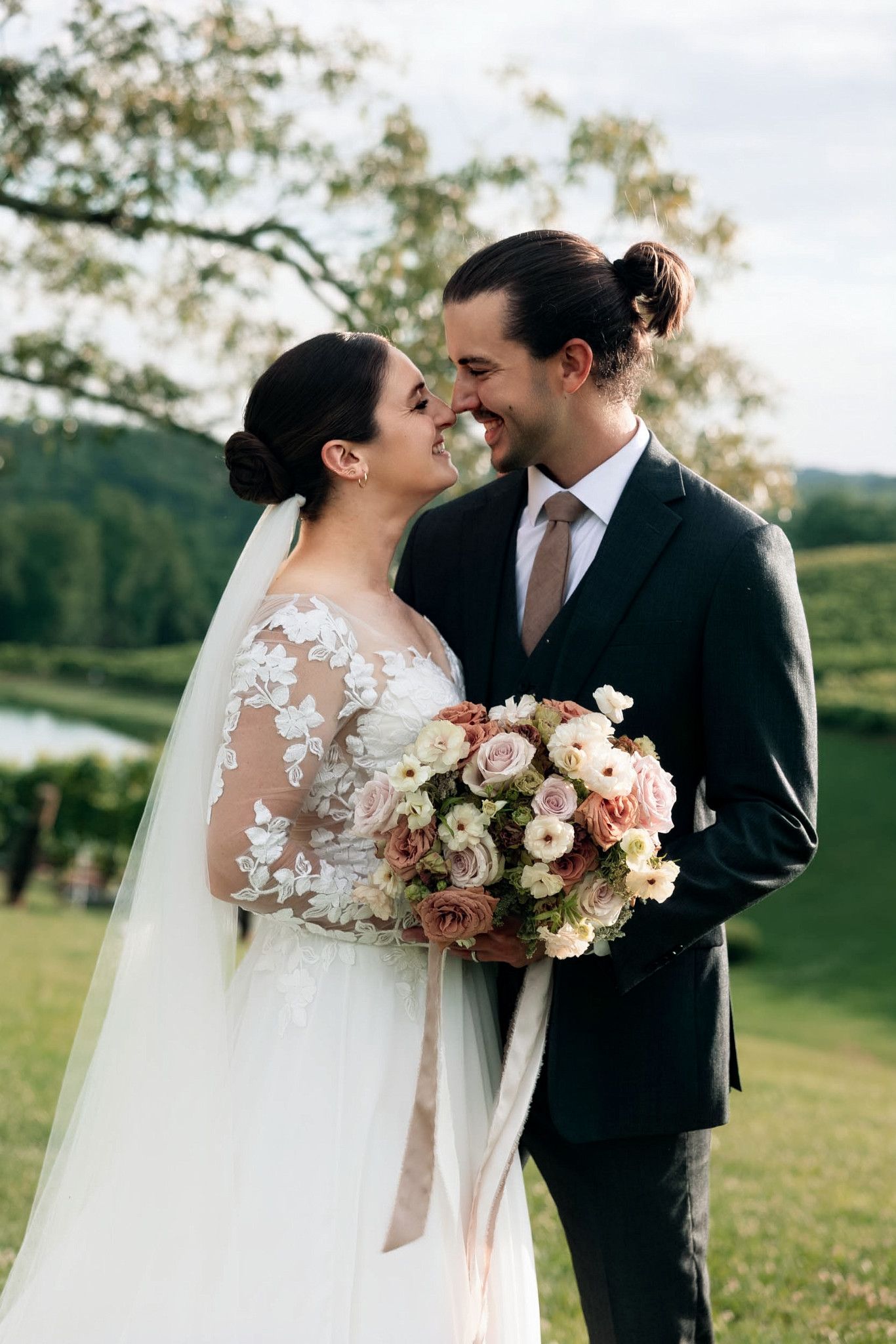 Bride and groom share a romantic moment outdoors, bride holding a blush floral bouquet in lace gown with veil.