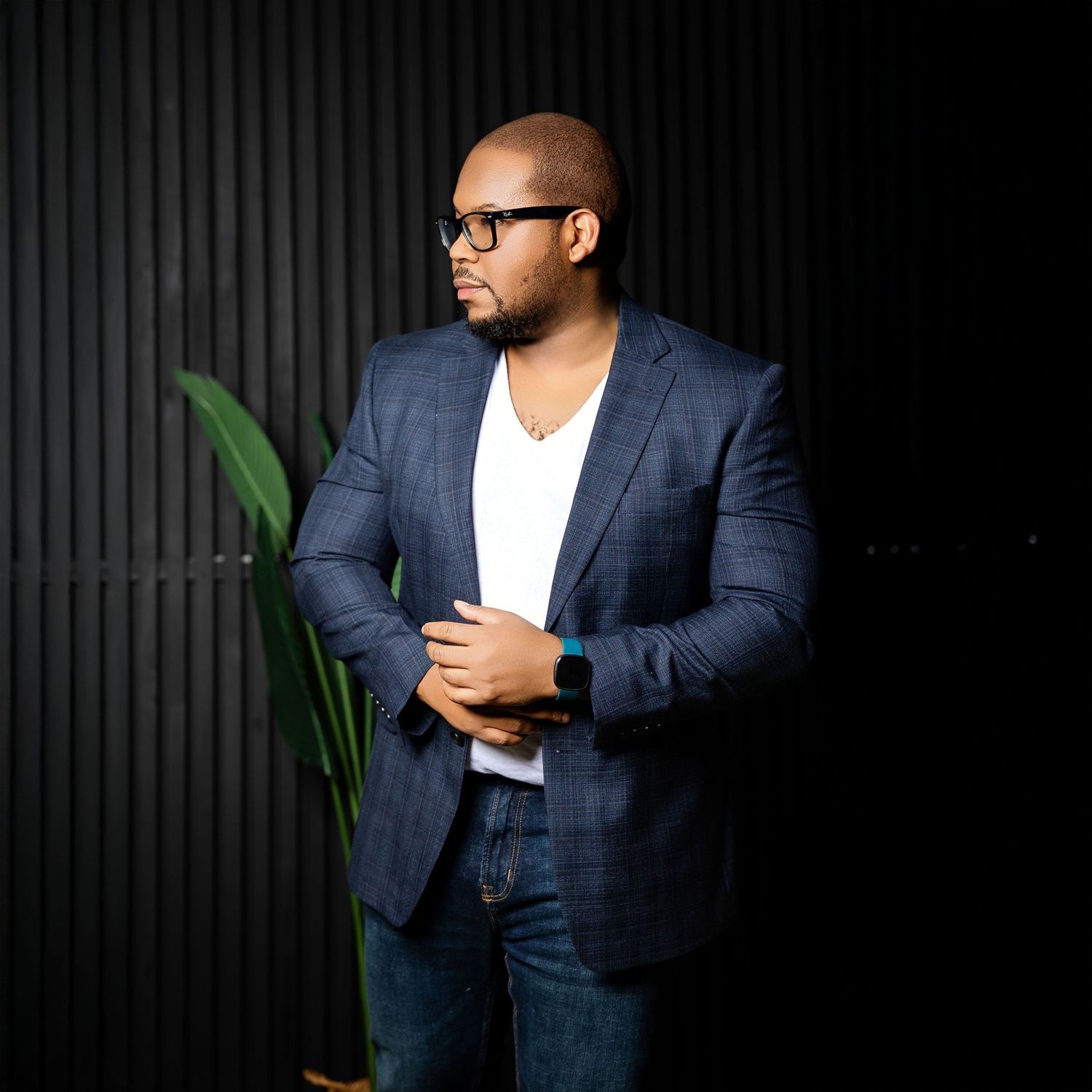A stylish man in a navy blazer and white shirt poses confidently against a dark textured wall with a plant nearby.