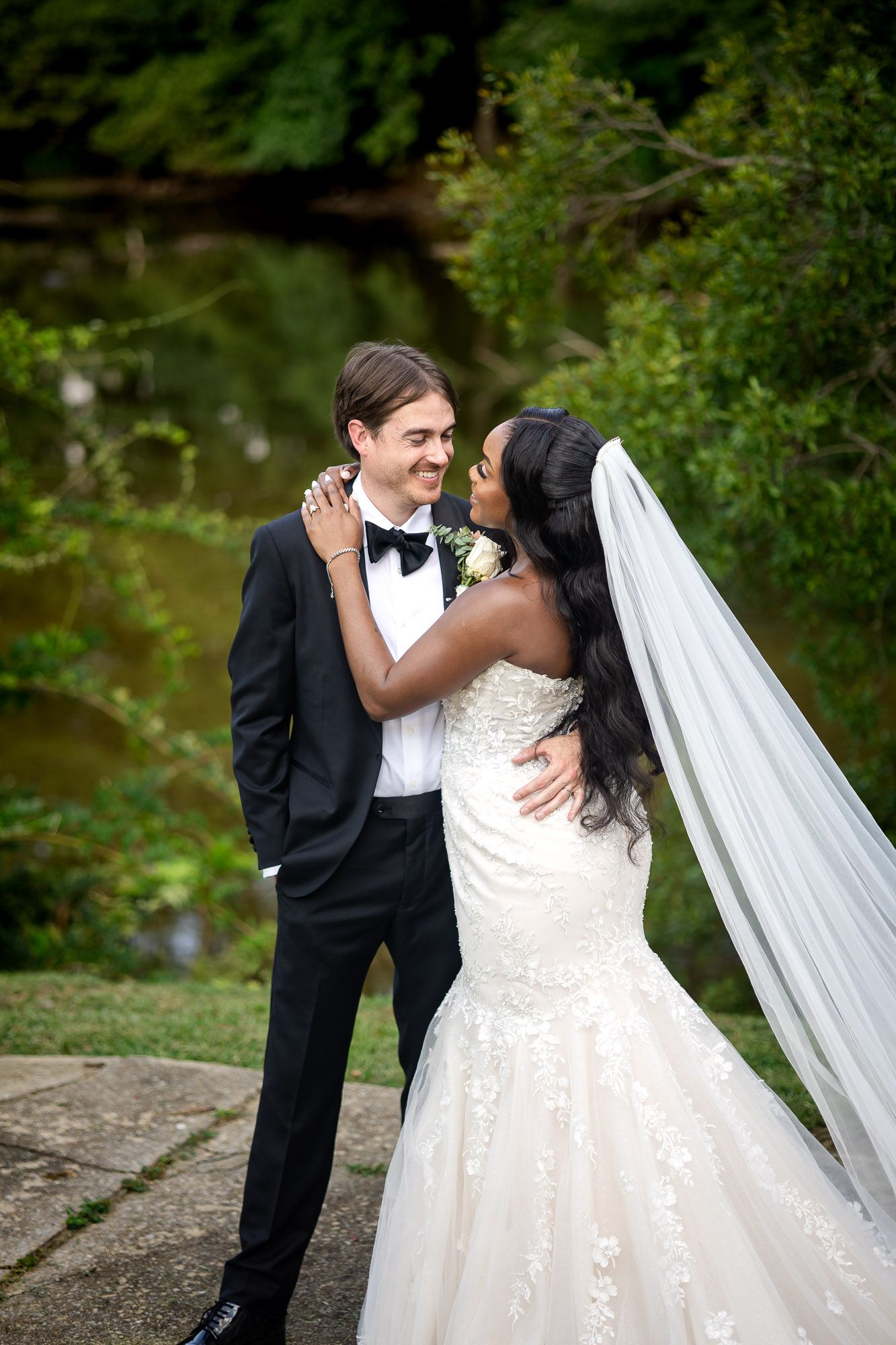 Interracial couple in wedding attire embracing outdoors, bride in lace gown with veil, groom in black tuxedo.