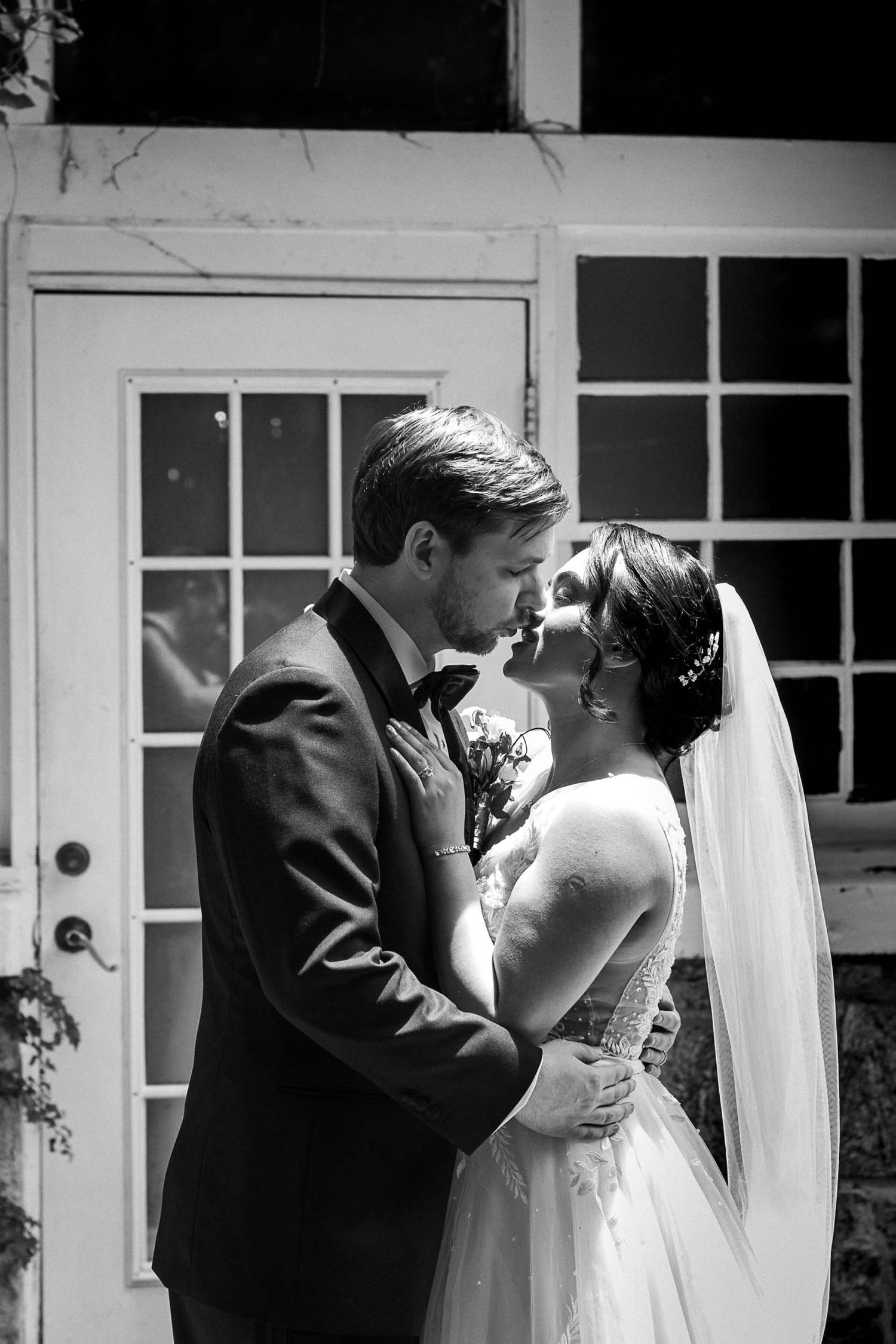 Black and white photo of a bride and groom sharing a tender moment, nearly kissing, outside a white building.
