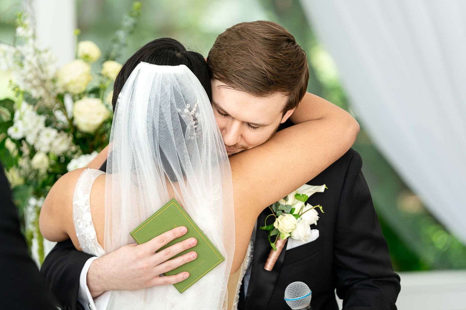Bride and groom share a heartfelt embrace during their wedding ceremony, with floral arrangements in the background.
