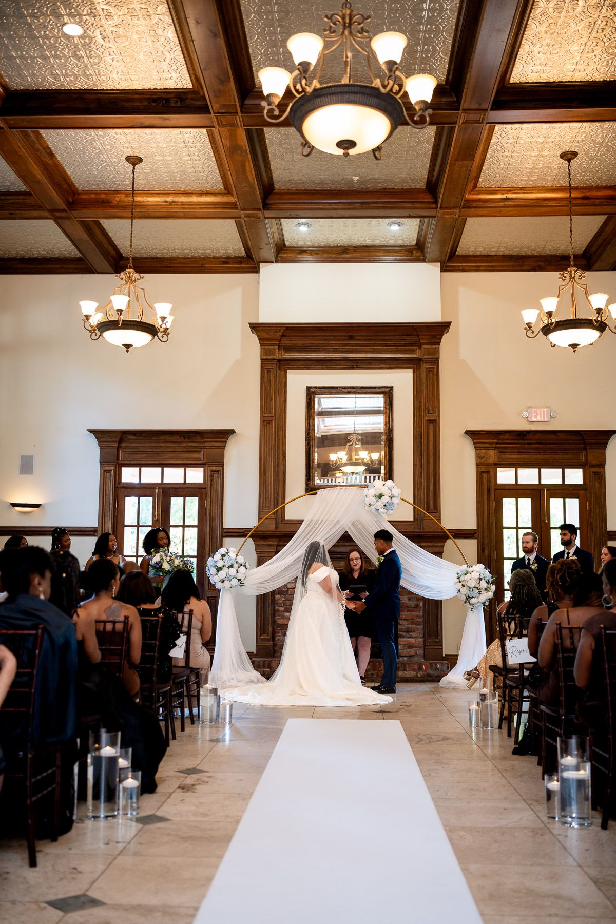 Elegant indoor wedding ceremony with couple at floral arch, white aisle runner, candles, and guests seated in decorated venue.