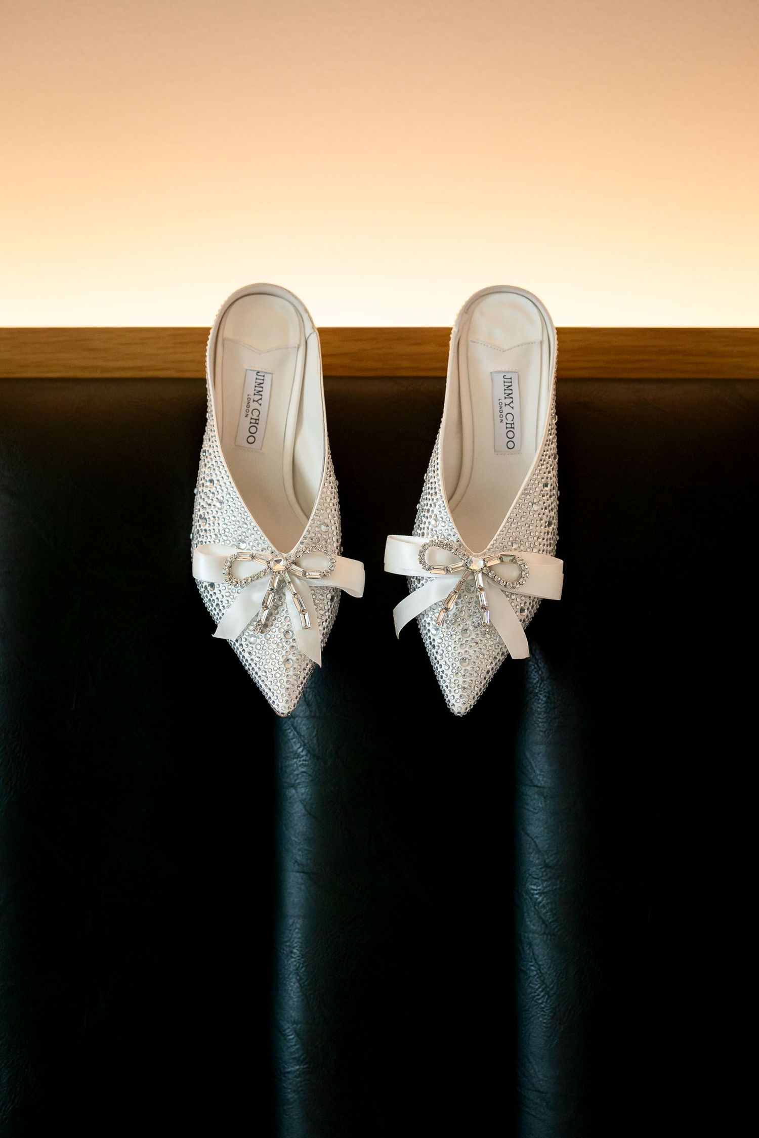 Elegant crystal-embellished Jimmy Choo bridal mules with satin bow detail on dark leather surface.