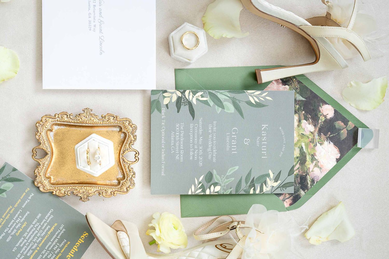 Elegant sage green wedding invitation suite with floral envelope liner, gold ring dish, and white heels on a marble surface.