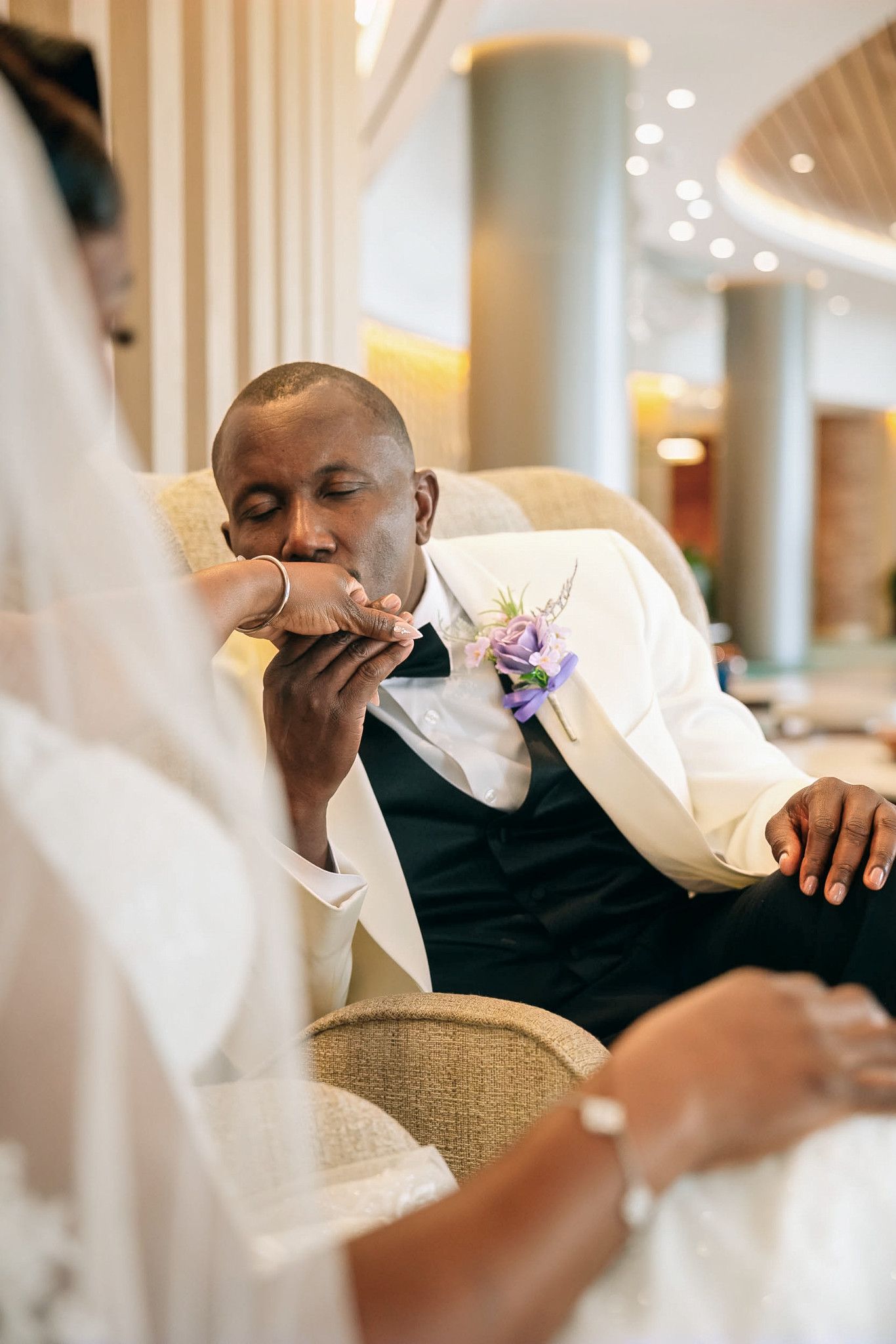 Groom in white tuxedo kissing bride's hand while seated in elegant chair during wedding ceremony.