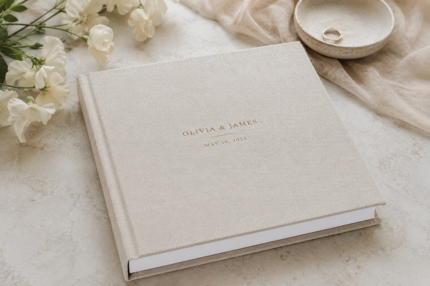 Elegant linen-covered wedding photo album with gold foil names on a marble surface with white flowers.