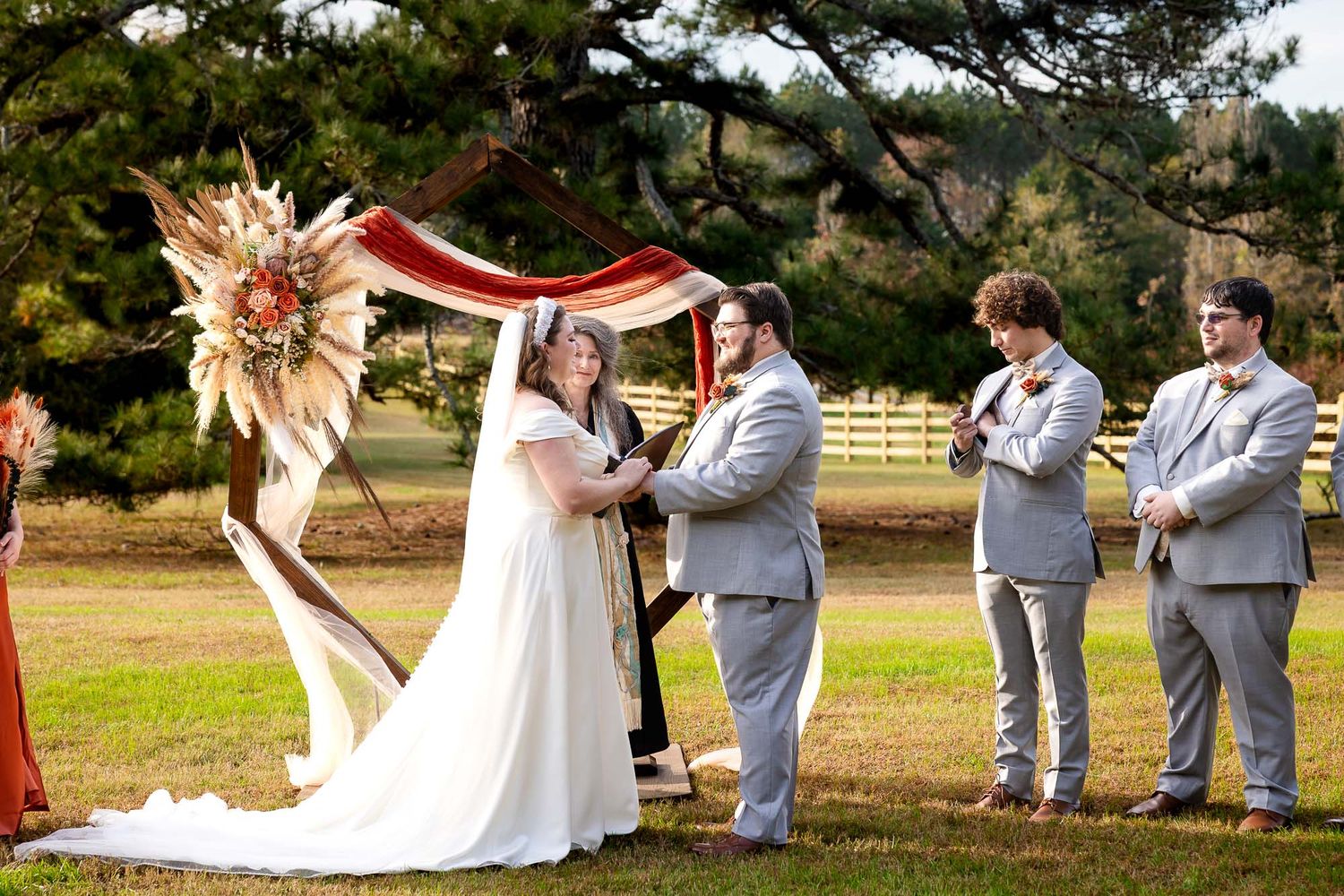 Outdoor wedding ceremony with bride and groom under floral arch, officiant and groomsmen in gray suits on green lawn.