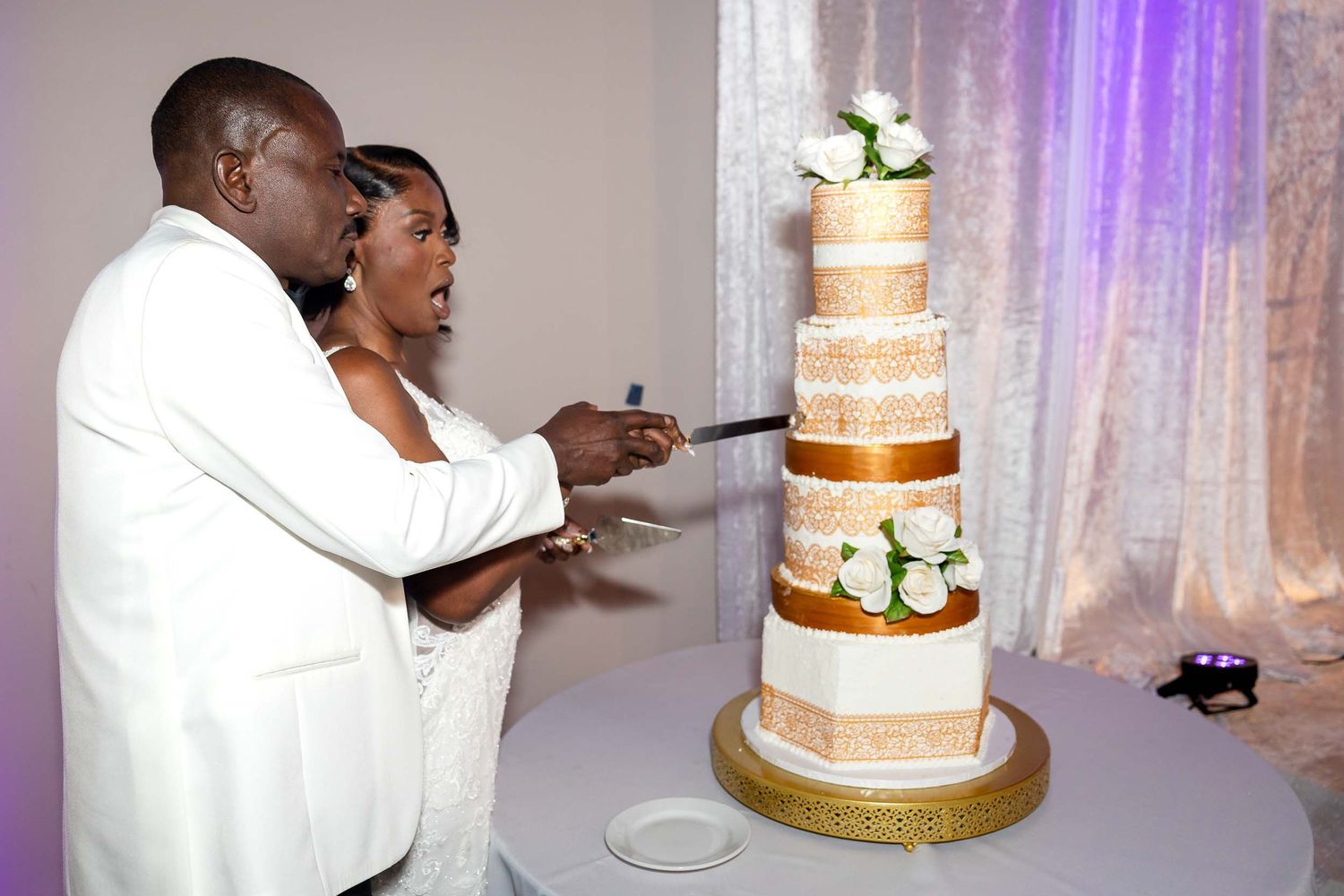 Bride and groom cutting a tall five-tier white and gold wedding cake decorated with white roses.