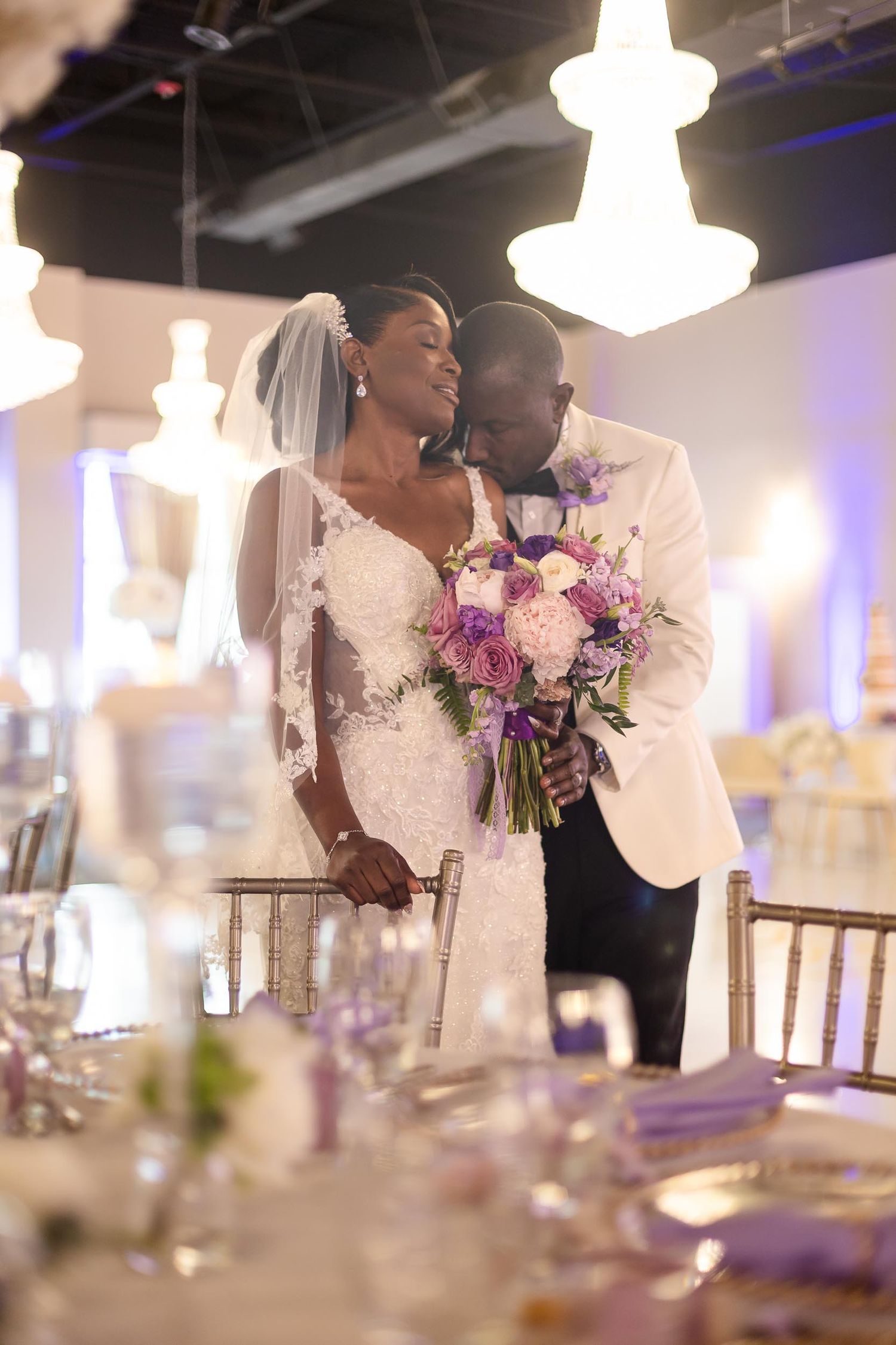 Bride and groom share romantic moment in decorated reception hall with chandelier lighting.