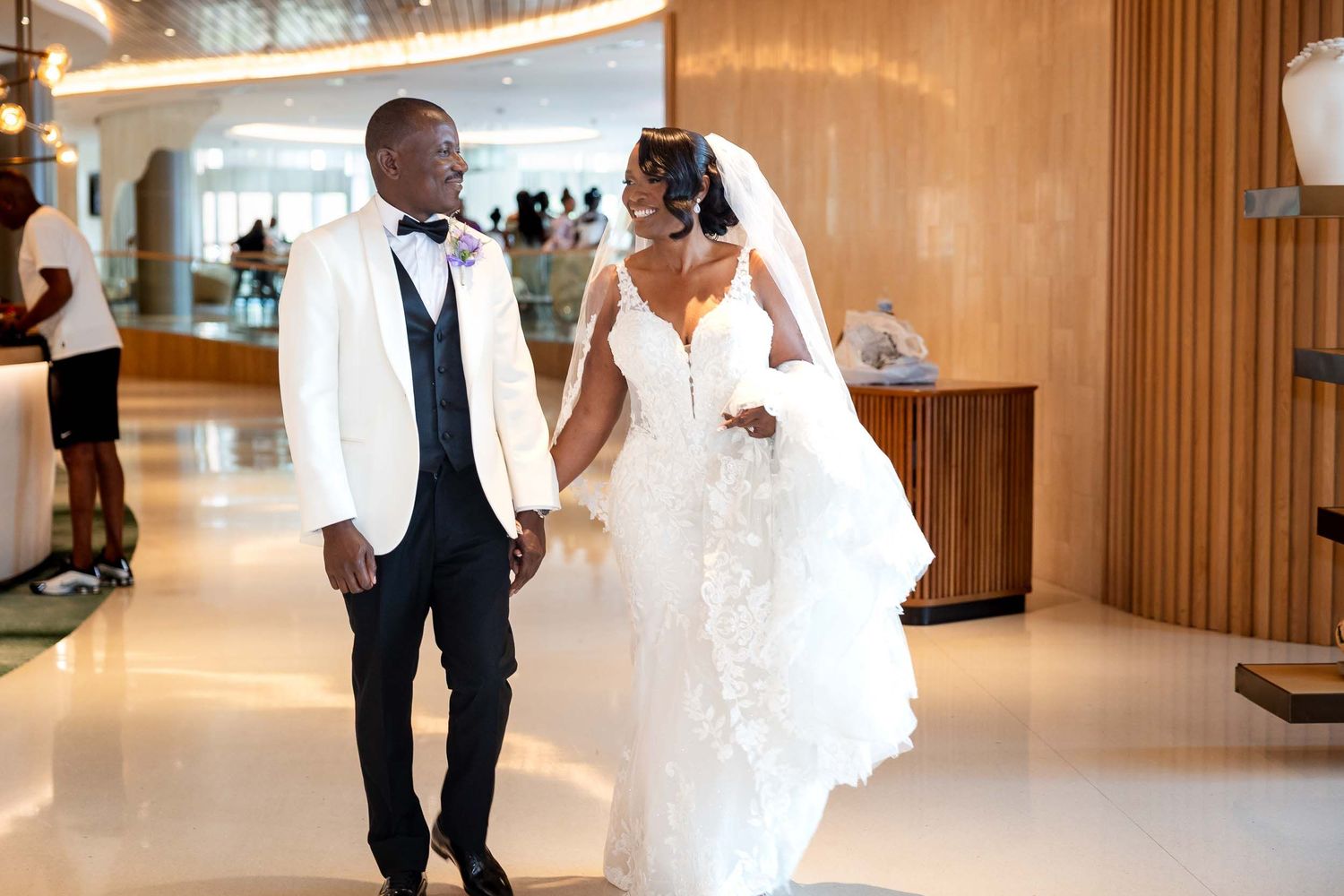 Elegant Black couple in wedding attire walking hand in hand in a modern venue, groom in white tuxedo, bride in lace gown.