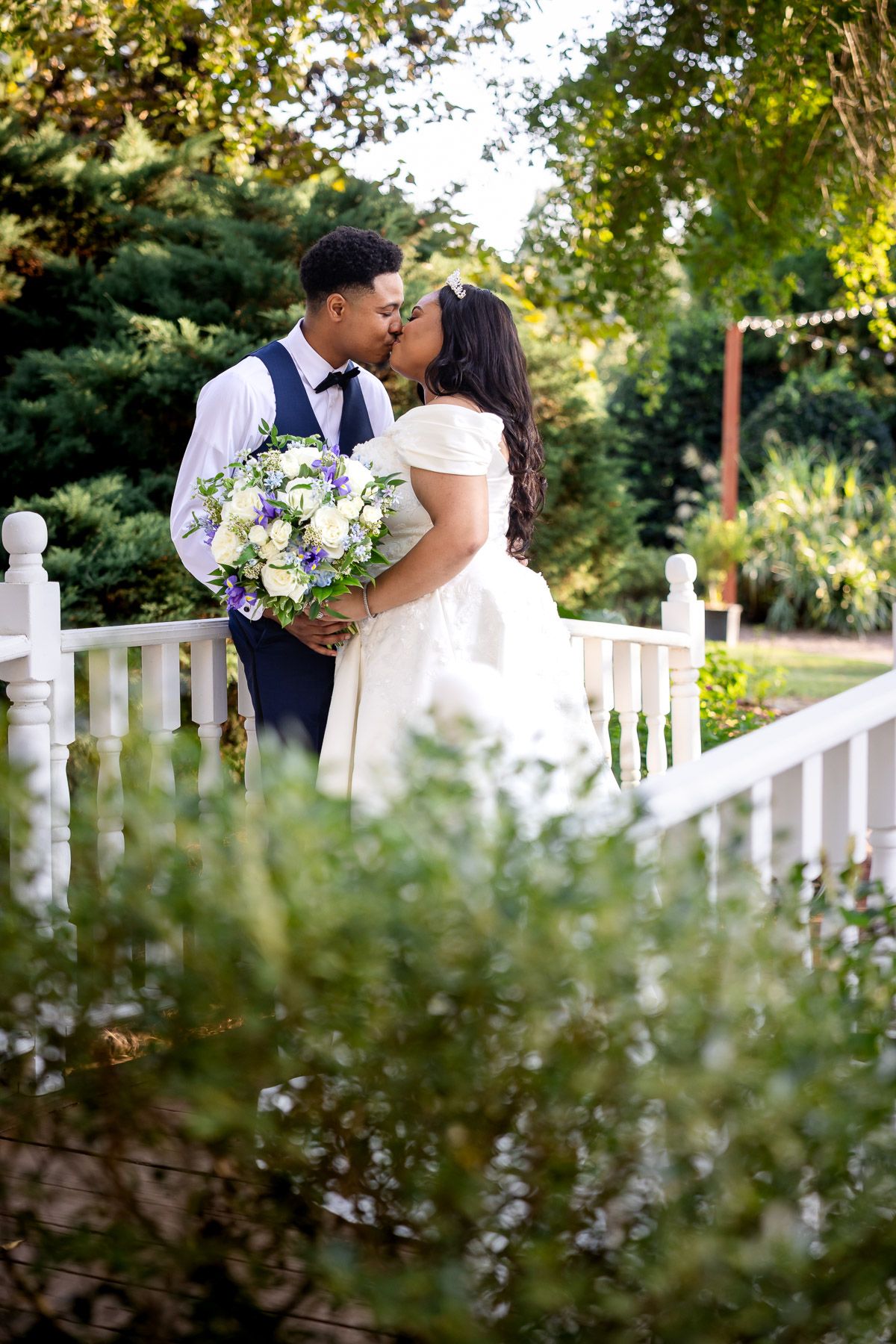 Bride and groom share a romantic kiss on a white garden bridge, bride holding a purple and white bouquet.