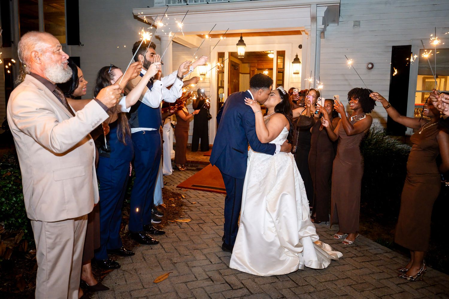 Bride and groom share a kiss during sparkler send-off exit surrounded by cheering wedding guests at night.