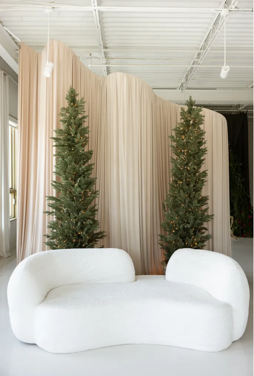 White curved sofa flanked by tall slim Christmas trees against flowing blush drape backdrop in bright modern space.