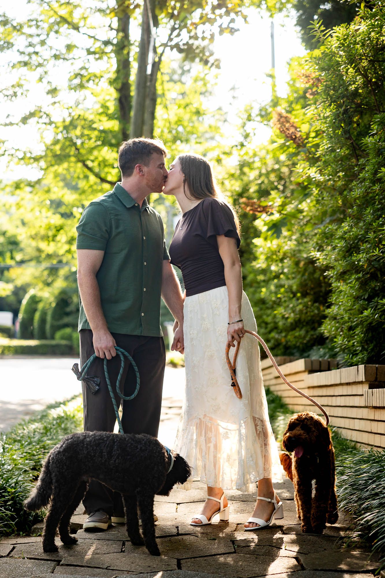 Couple kissing while walking two dogs, a black poodle and brown doodle, on a sunny garden path surrounded by lush greenery.