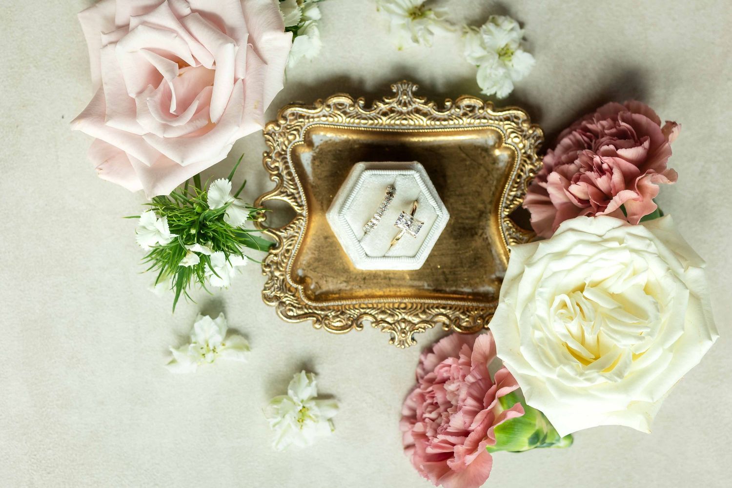 Gold ornate tray with wedding rings in velvet box surrounded by pink, white, and mauve roses and carnations on neutral background.