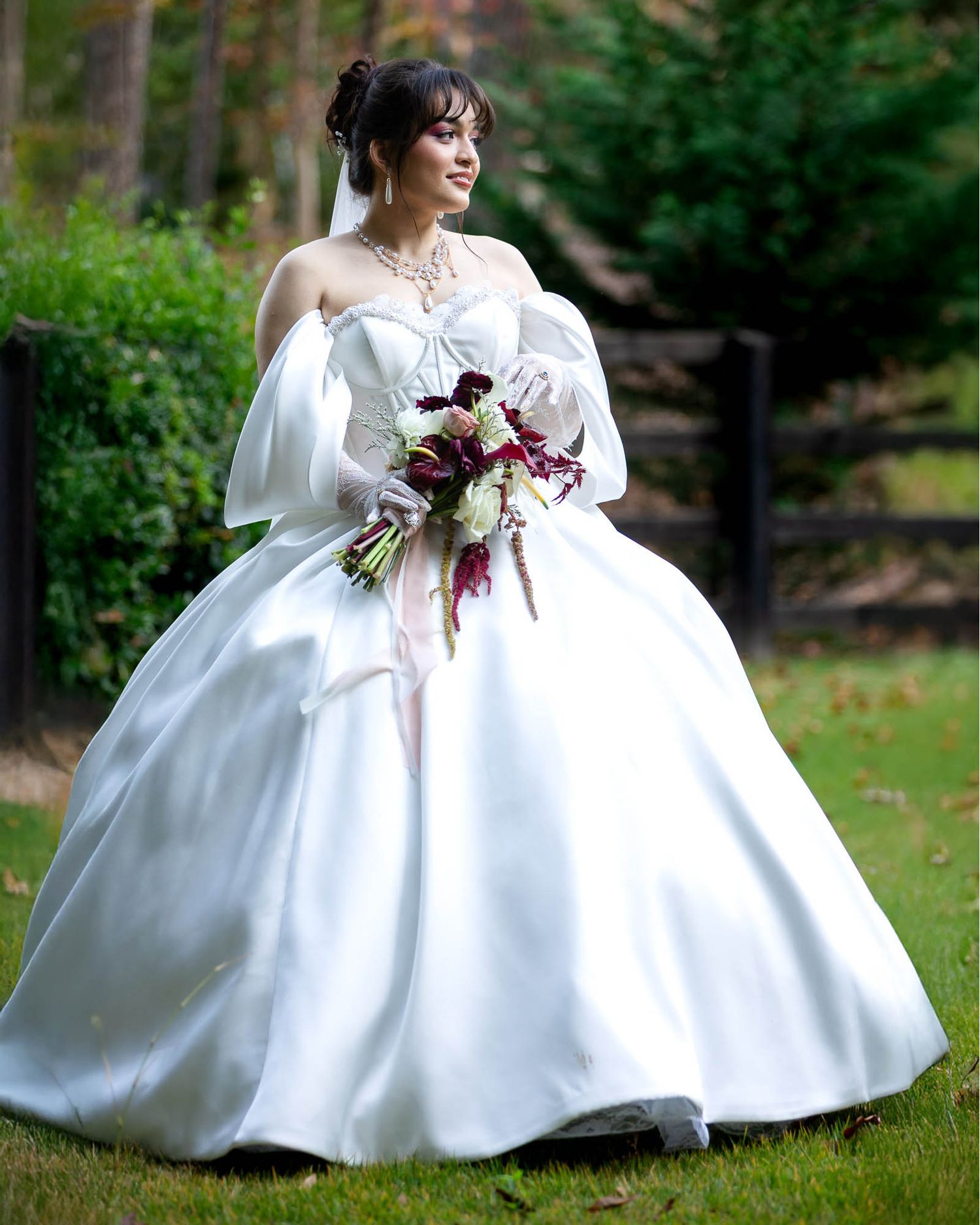 Bride in a white ball gown with off-shoulder sleeves holds a burgundy and white bouquet outdoors.