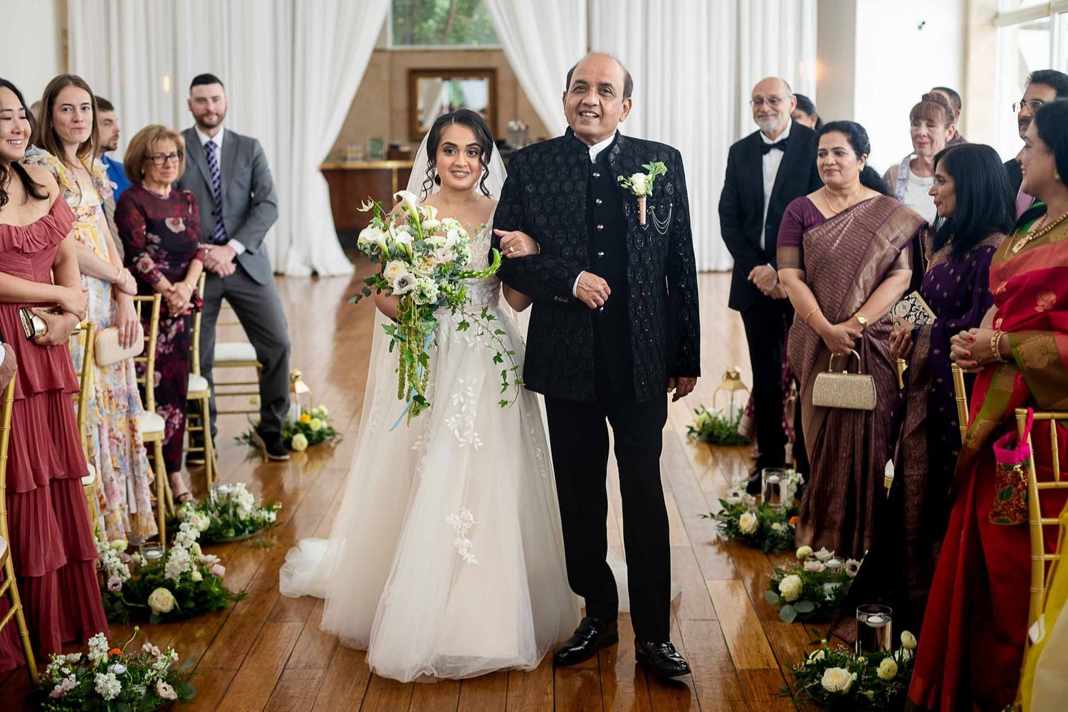 Bride in white gown walks down aisle with father in black suit at elegant indoor wedding ceremony.