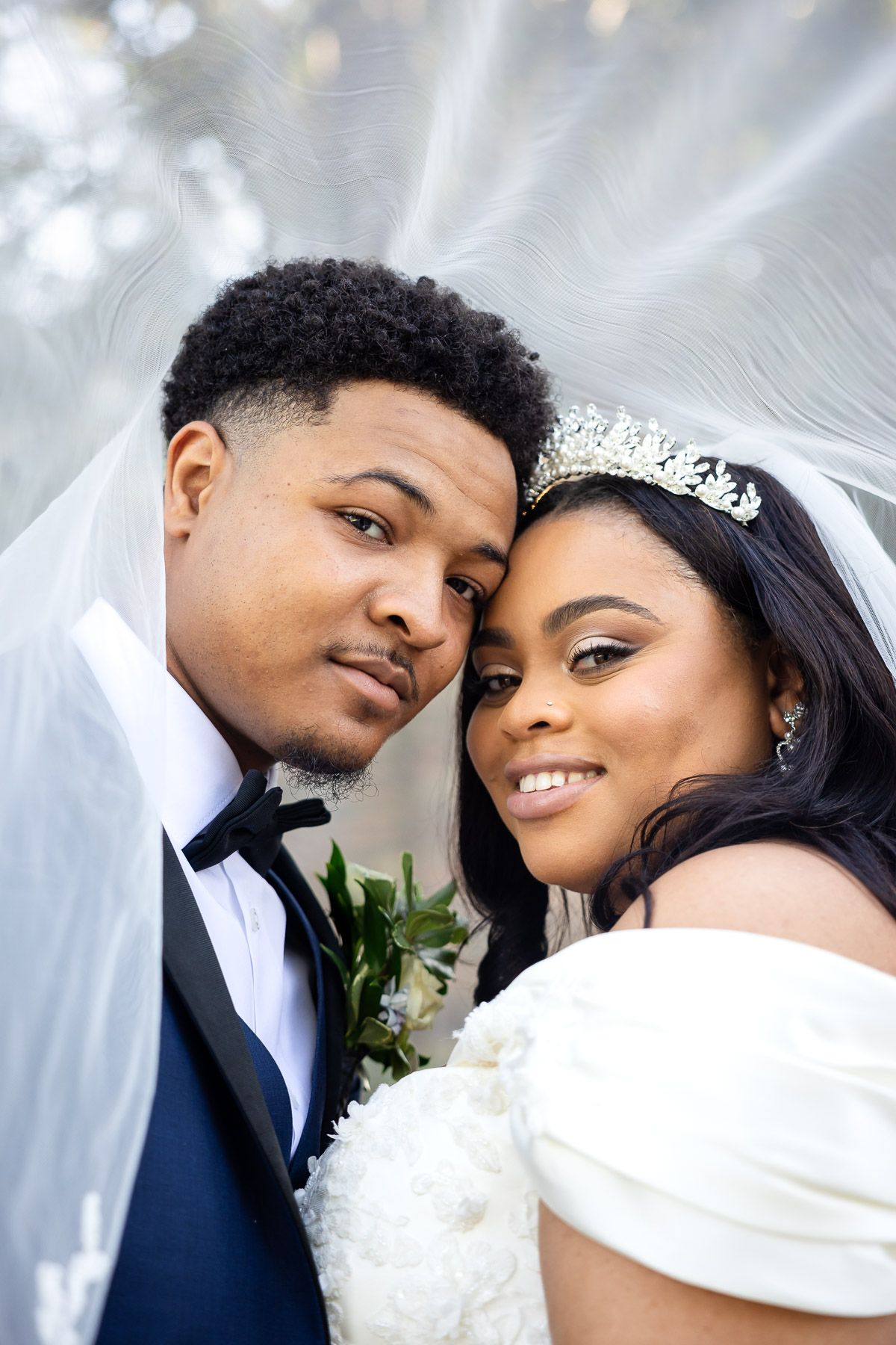 A bride and groom pose closely together under a flowing veil, the bride wearing a tiara and white gown on their wedding day.