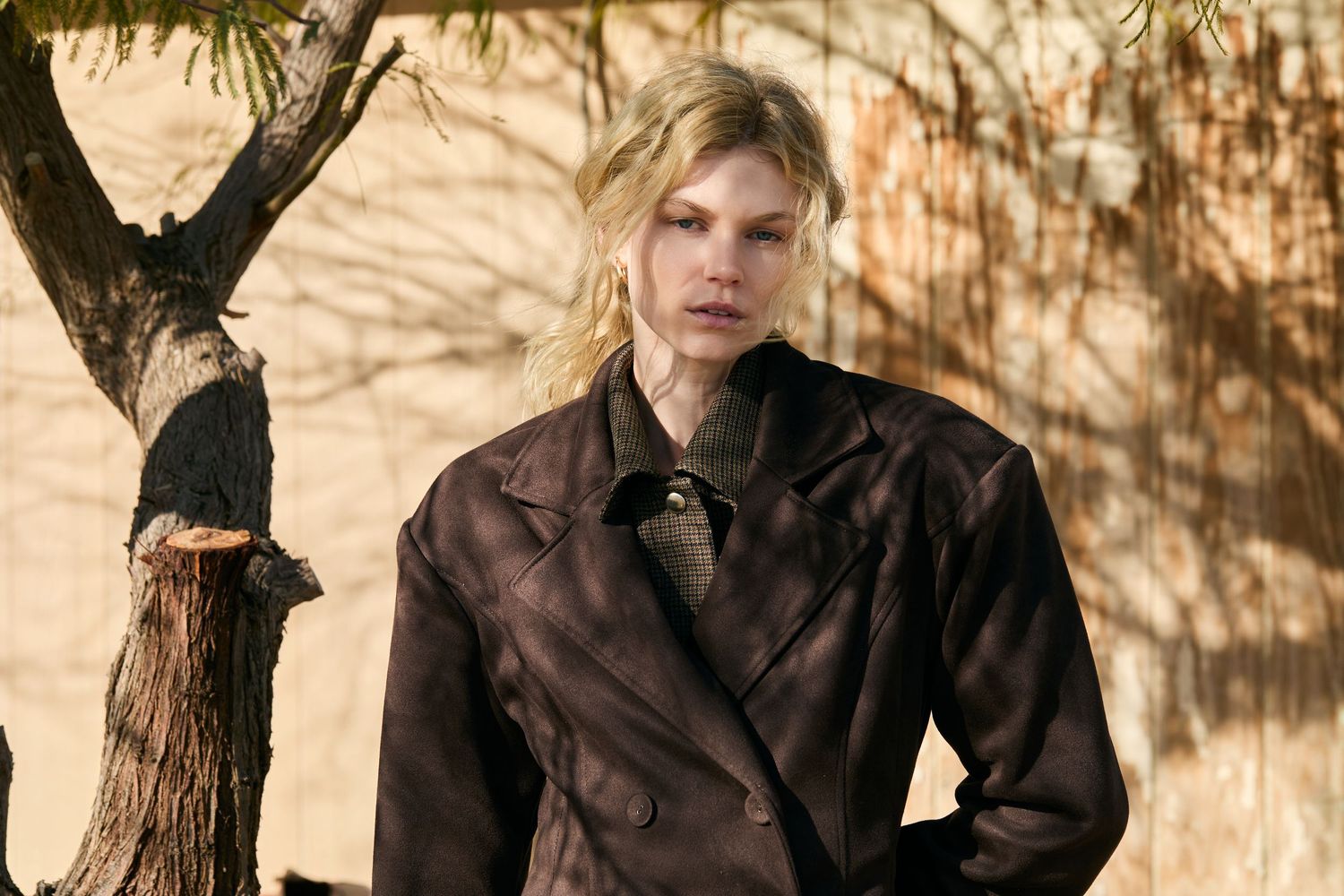 Person in brown coat posing outdoors against textured wall with tree shadows.