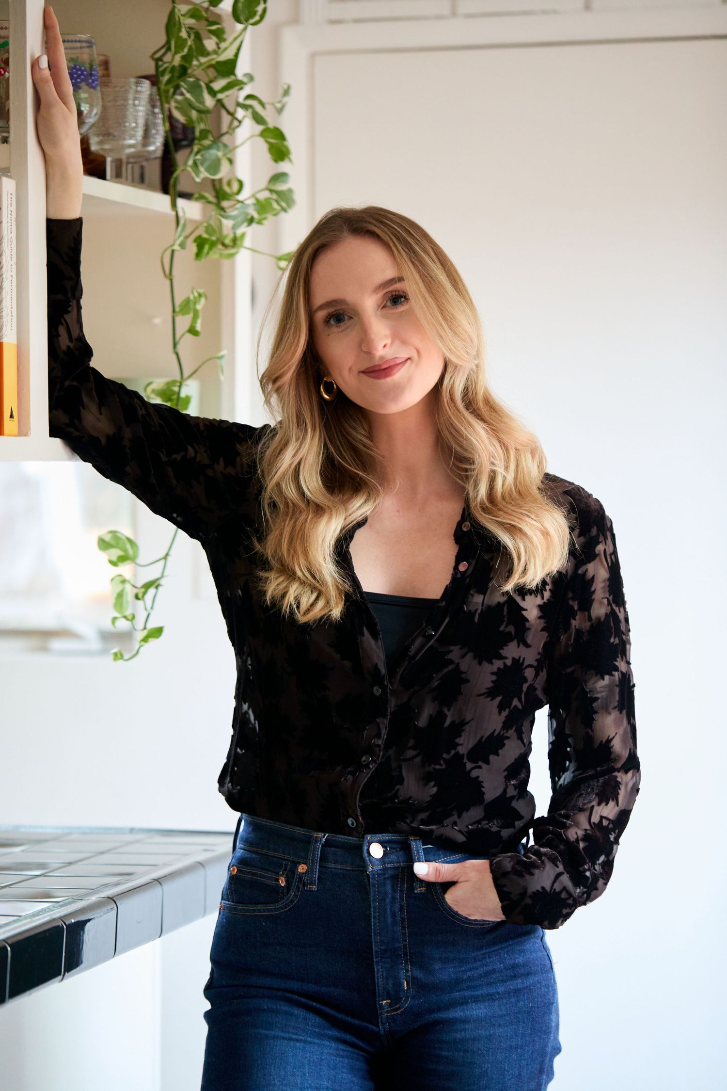 Blonde woman in black floral blouse and jeans posing by a white shelf with plants in a bright modern kitchen.