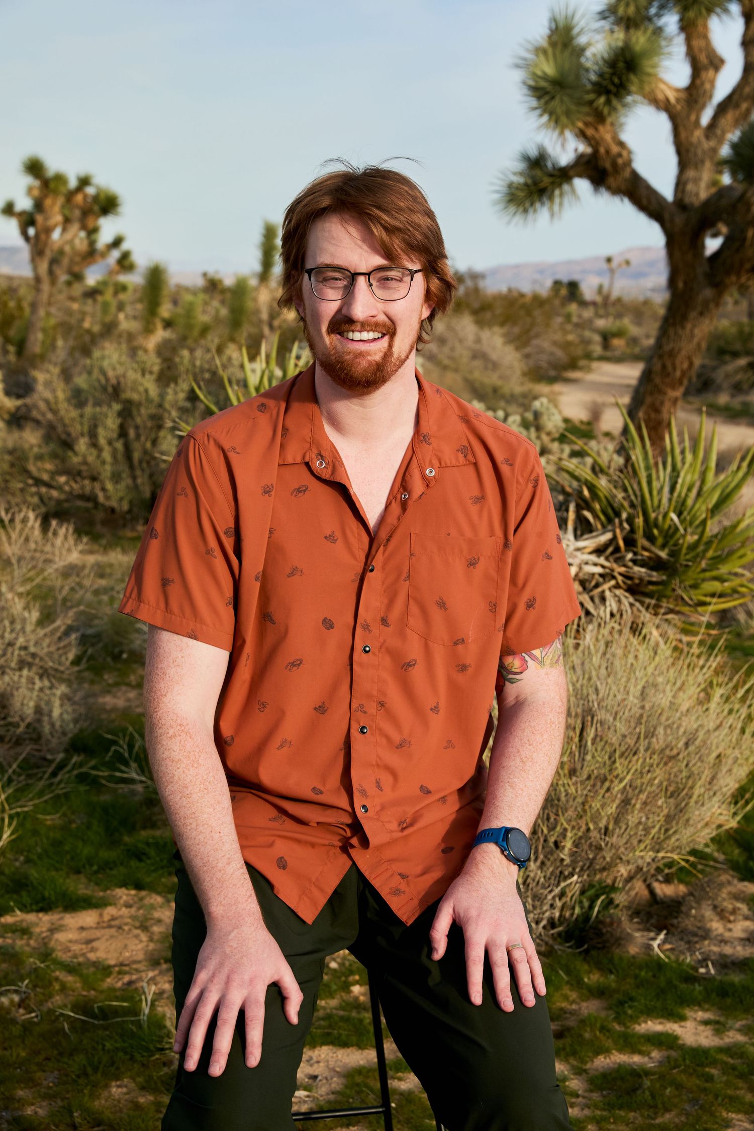 A person in a rust-colored button down shirt poses outdoors in a desert landscape with Joshua trees in the background.