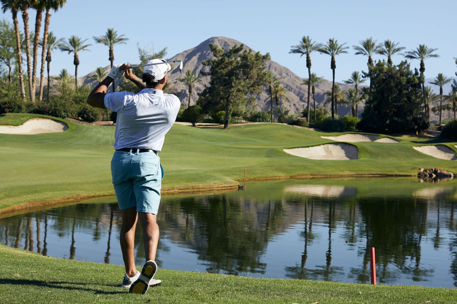 Golfer taking a swing on a scenic course with palm trees and mountains in the background.