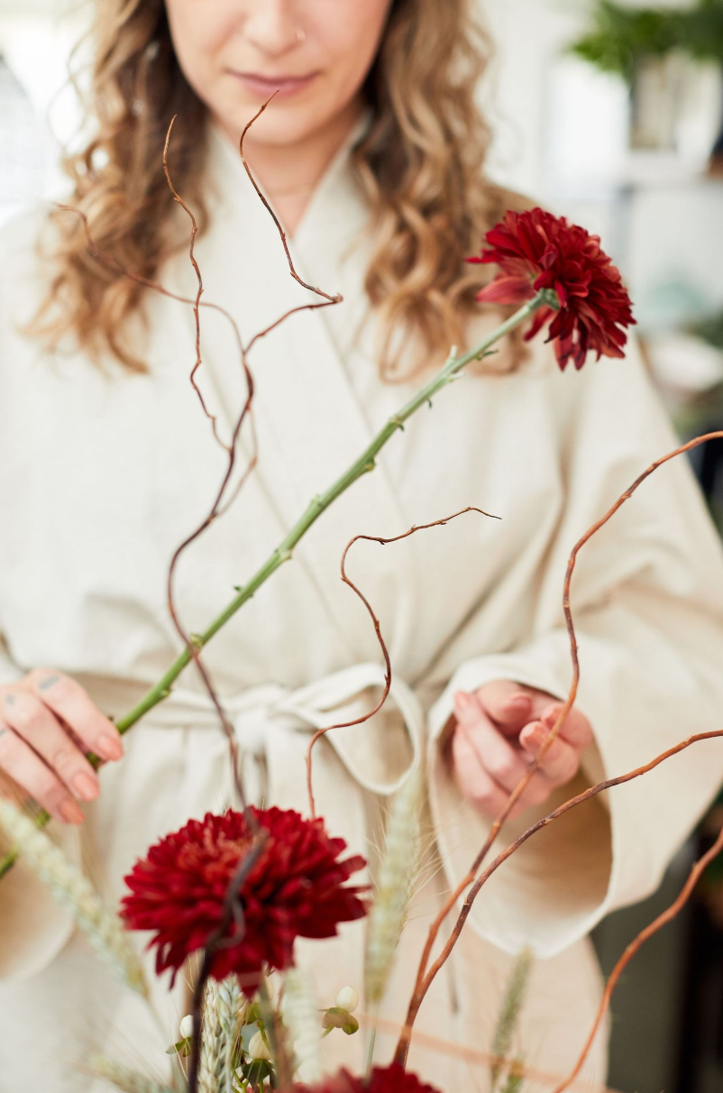 Person in cream colored garment arranging red carnation flowers with curvy branches in artistic display.