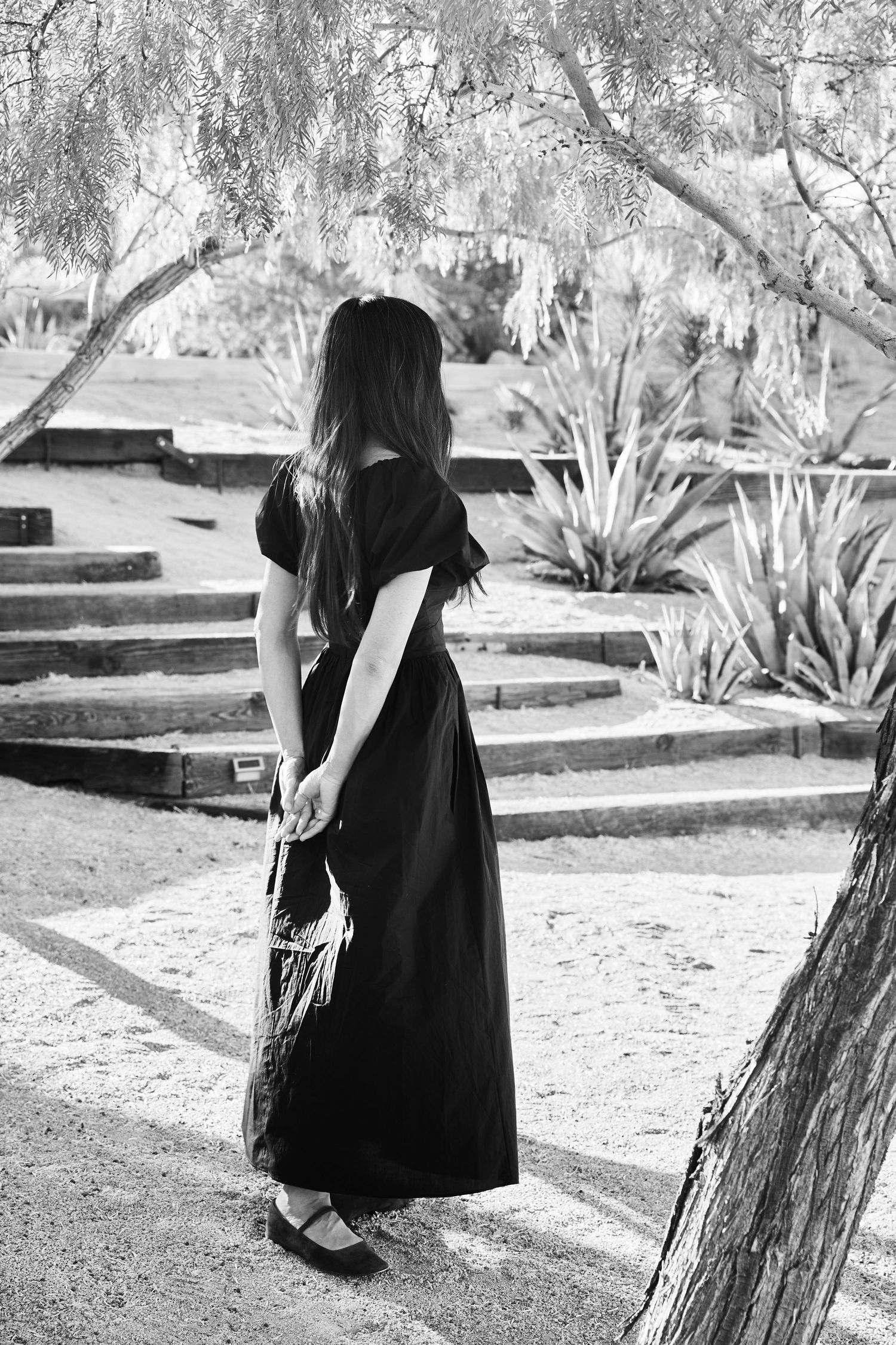 Black and white photo of a figure in a long dark dress standing on garden steps among desert plants and trees.