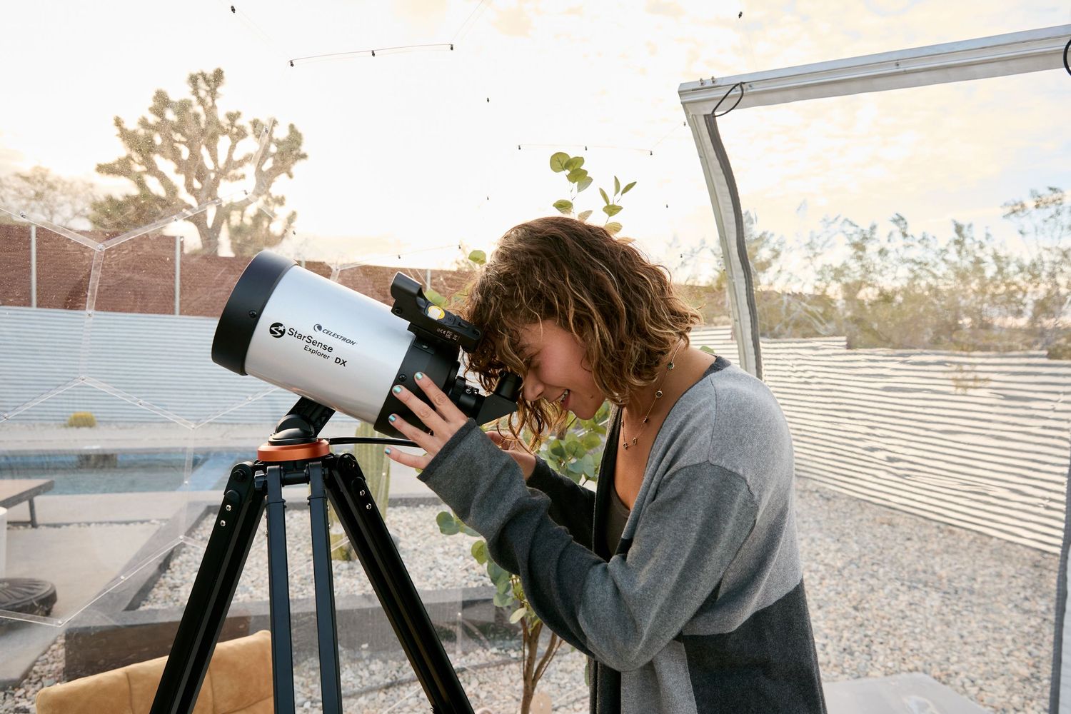 Woman looking through a Celestron telescope on an outdoor patio at sunset near Joshua trees.
