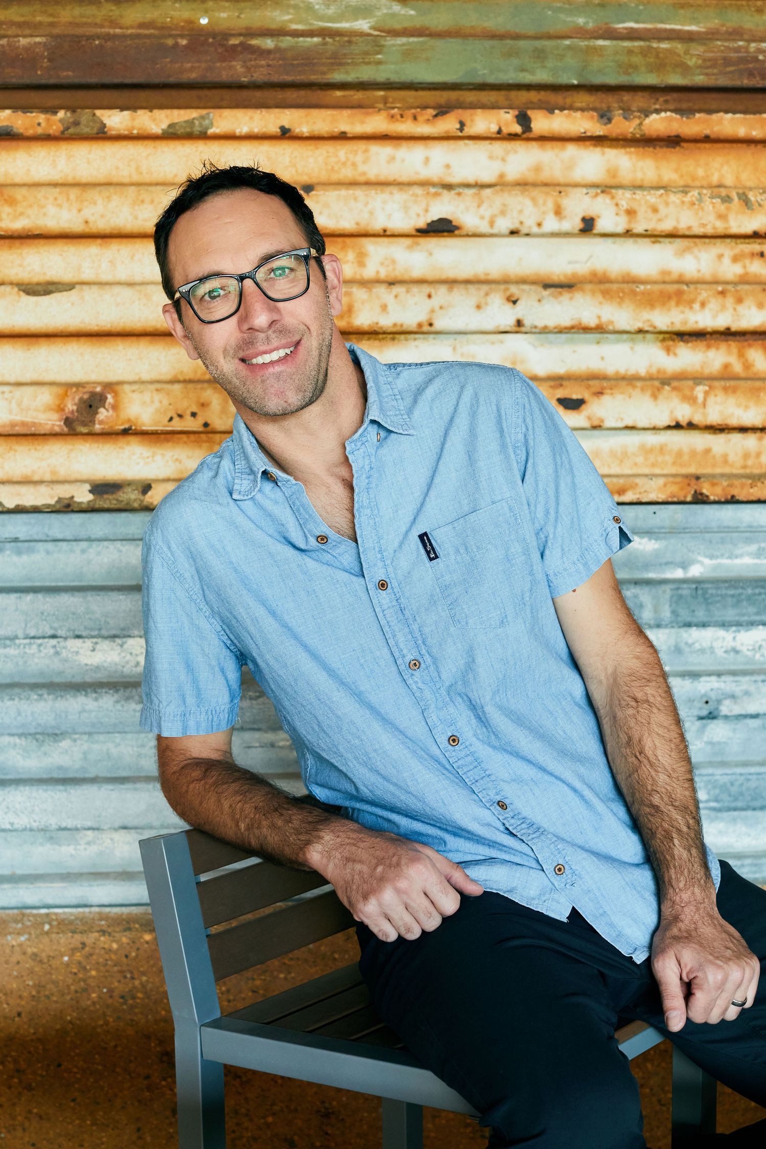 Man in blue shirt smiling while seated against rustic metal wall.