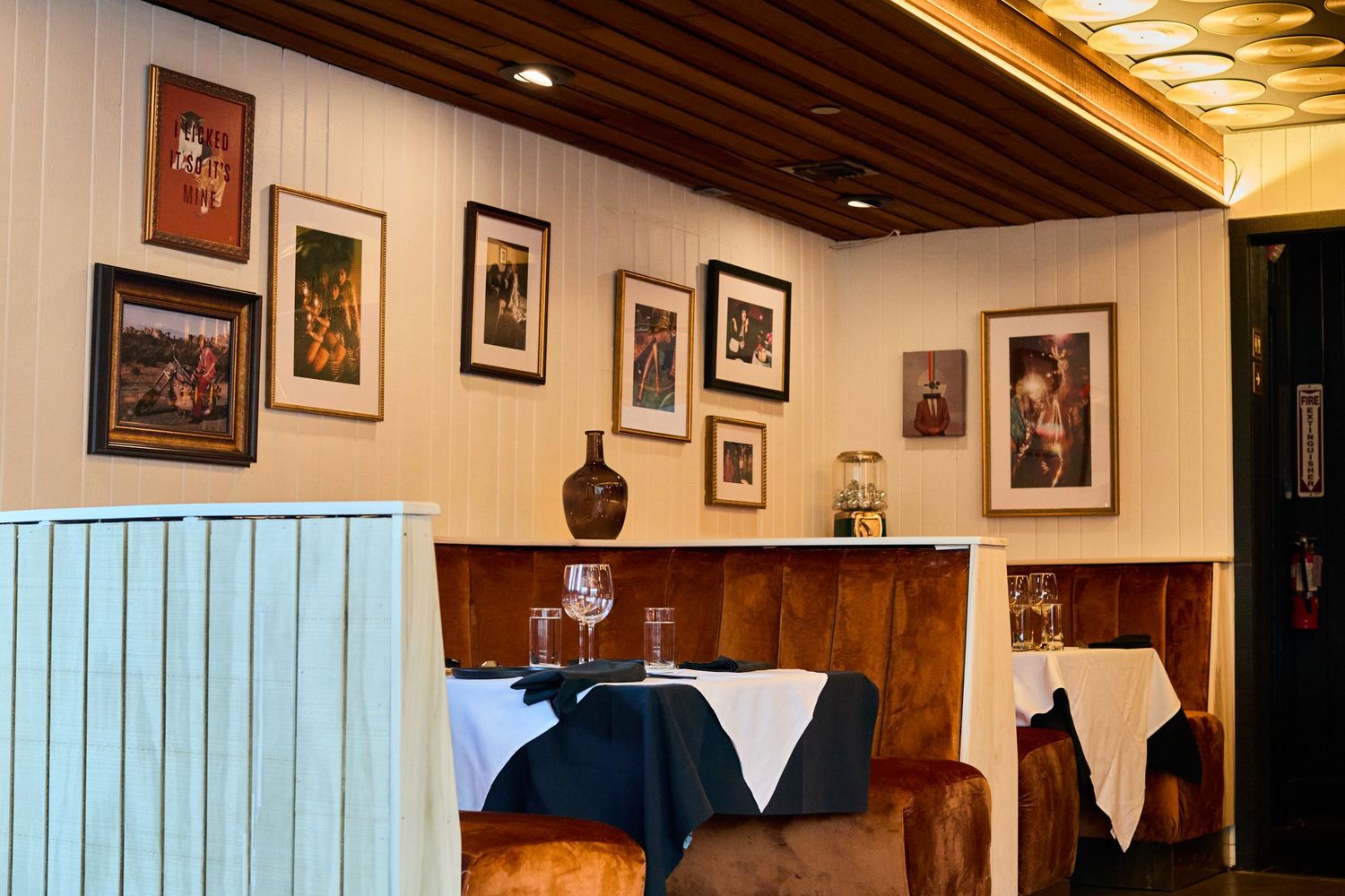 A rustic restaurant interior with framed photographs on white walls and dark wood paneling features white tablecloths and warm lighting.
