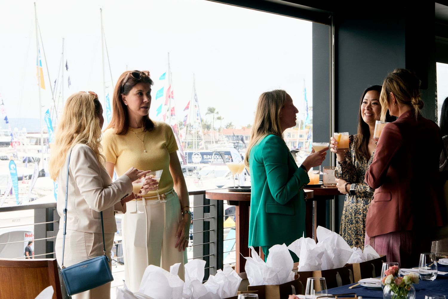 People enjoying drinks and conversation on a waterfront balcony with boats visible in the marina background.