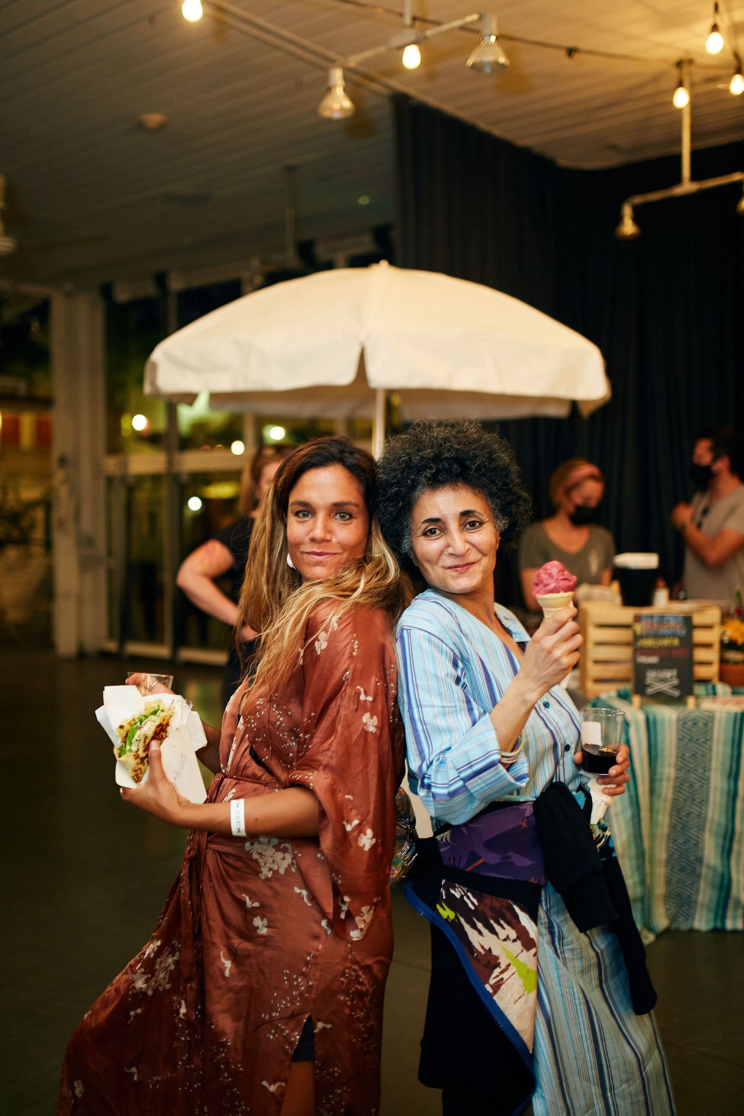 Two people enjoying food and ice cream beneath a white umbrella at an indoor event with string lights overhead.