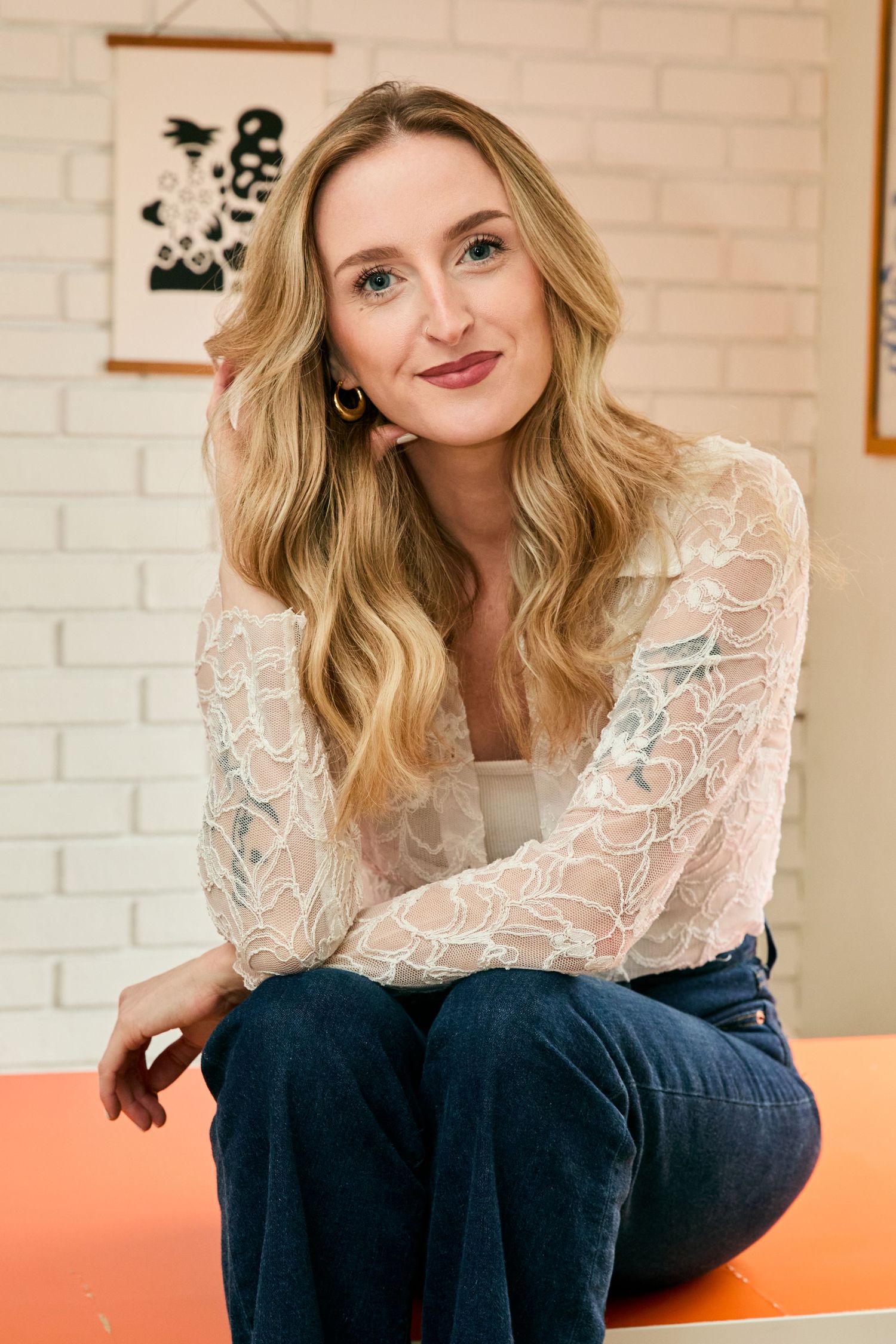 Blonde woman in white lace top and jeans sits relaxed, smiling softly in a bright white brick room.