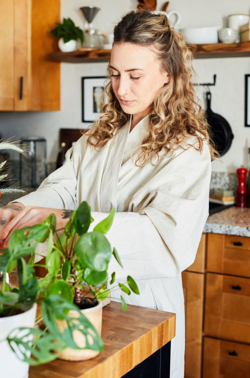 Person in white robe tending to houseplants in modern kitchen interior.
