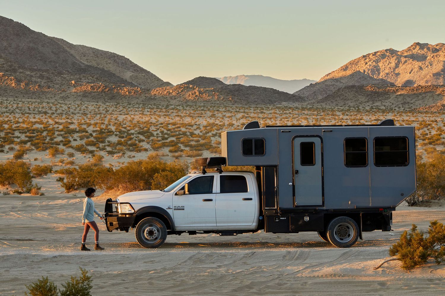 White truck with attached camper parked in desert landscape at sunset.