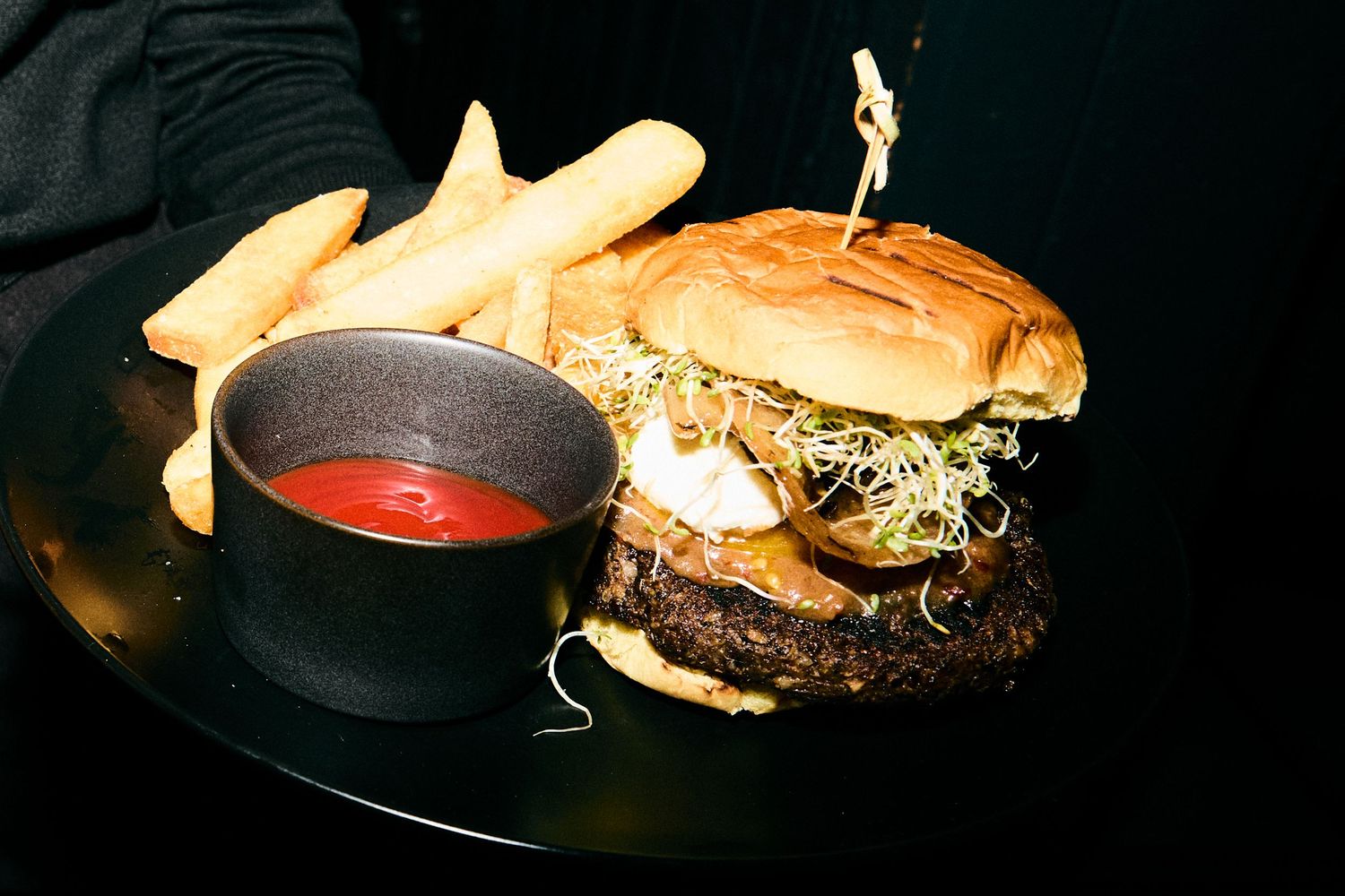 Juicy cheeseburger with golden french fries and red dipping sauce served on black plate against dark background.